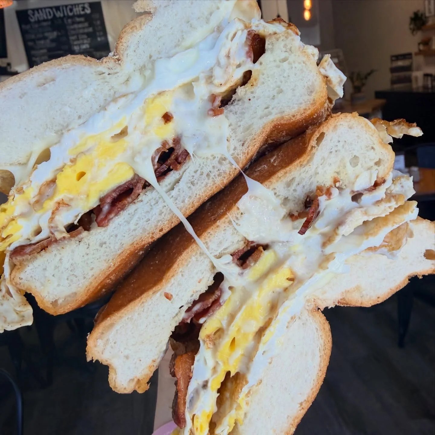 Close-up of a breakfast sandwich with fried egg, bacon, and bread, held in a person's hand inside a café.