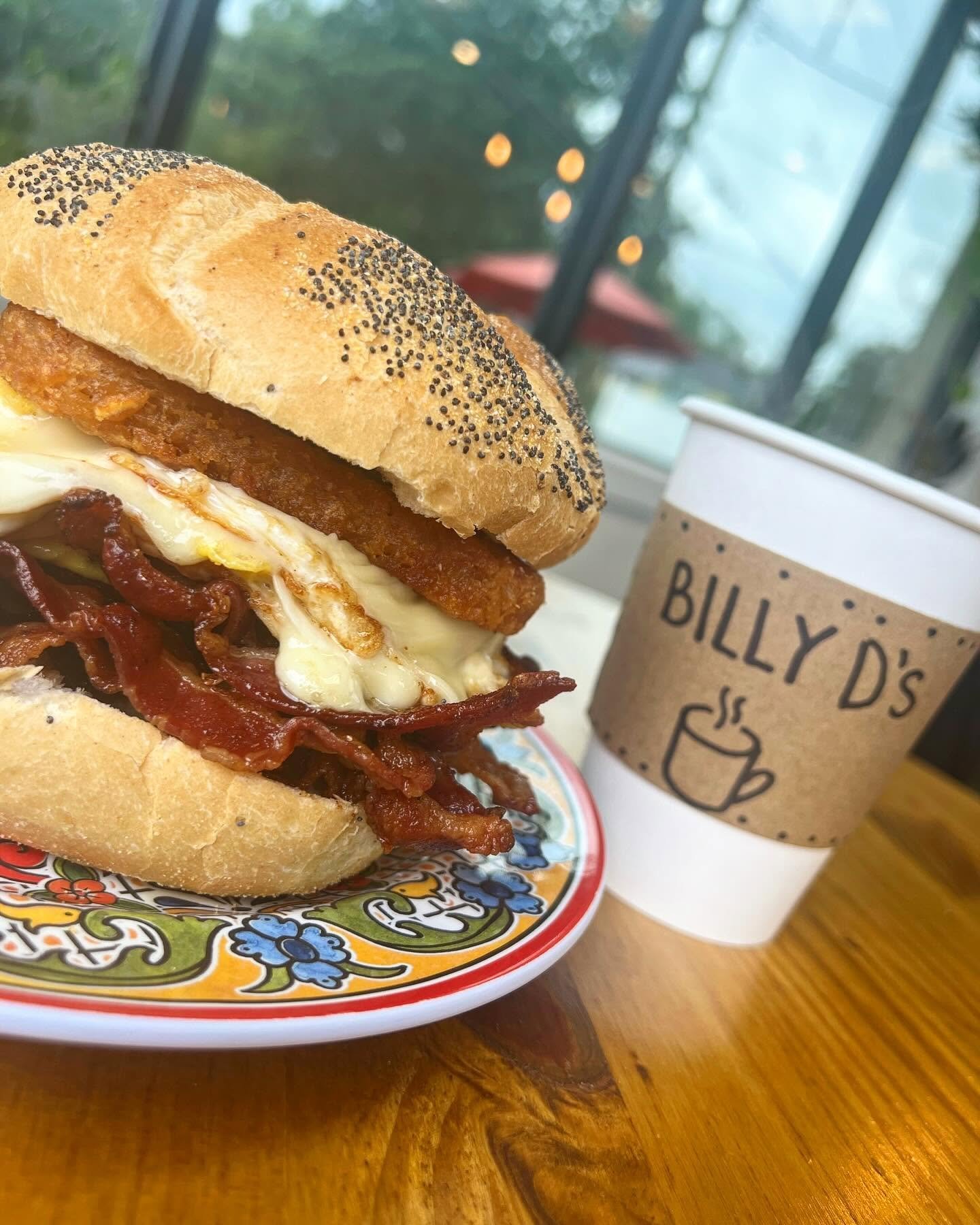 Close-up of a breakfast sandwich with fried egg, bacon, and hash brown on a poppy seed bun on a decorative plate. In the background, there is a to-go coffee cup from Billy D's with a stylized coffee cup logo and text, and a window showing outdoor sce