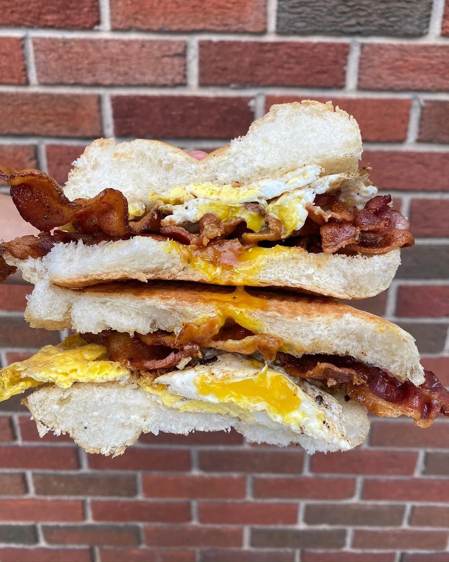 Close-up of a stack of sandwiches with bacon and fried eggs, set against a red brick wall.