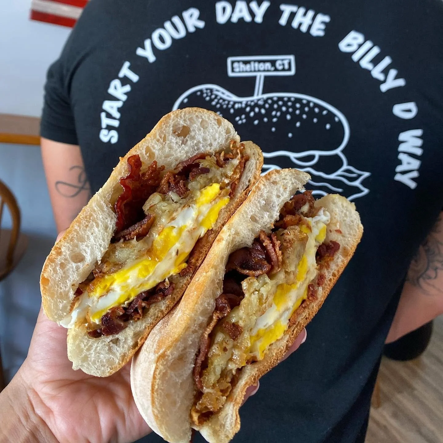 A person holding a sandwich with fried bacon, cheese, and a fried egg on a toasted bun, with another person wearing a black t-shirt with white text and illustration in the background.