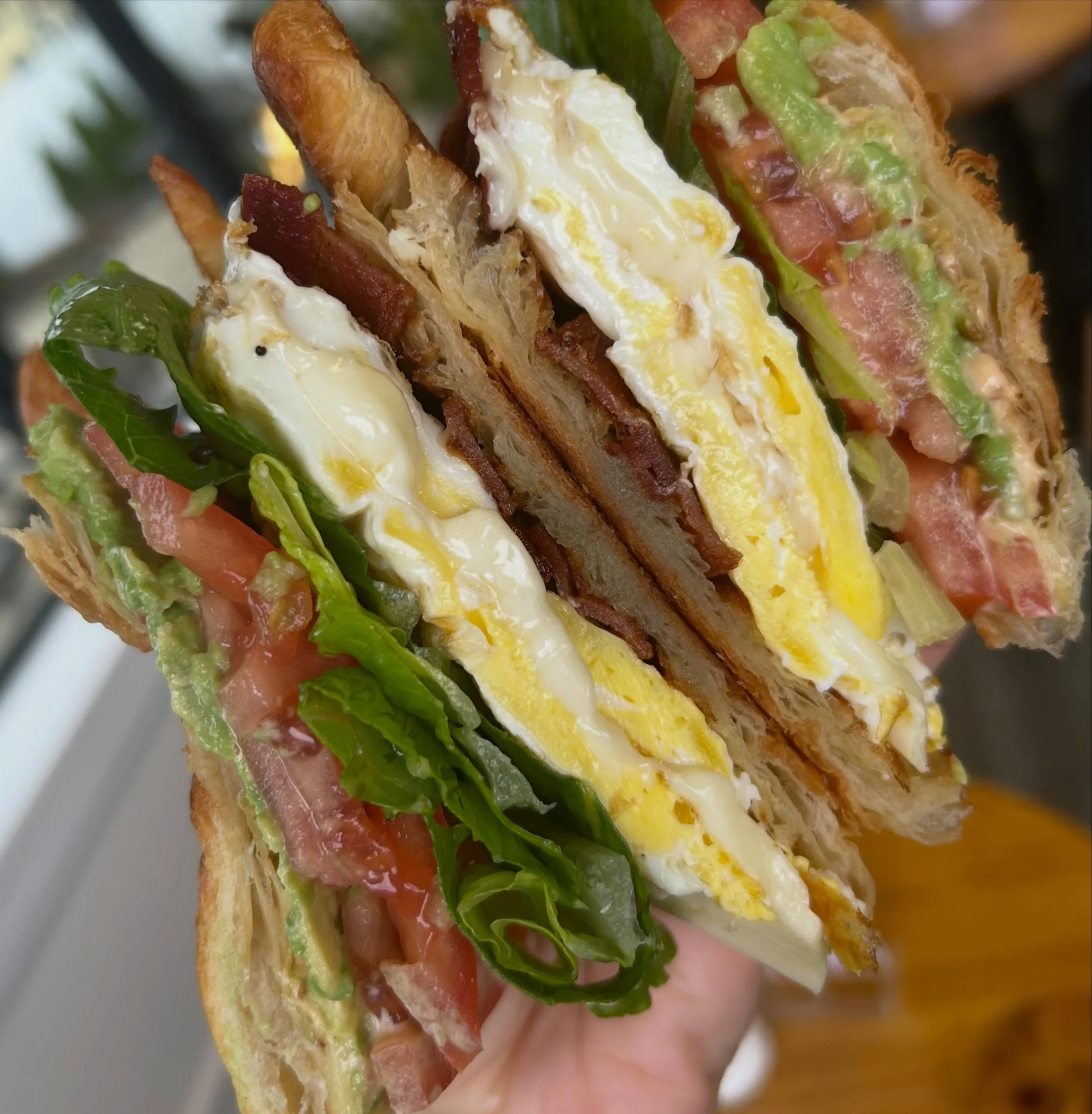 Close-up of a sandwich with layers of lettuce, tomato, slices of ham, scrambled eggs, cheese, and guacamole in a toasted bread bun.