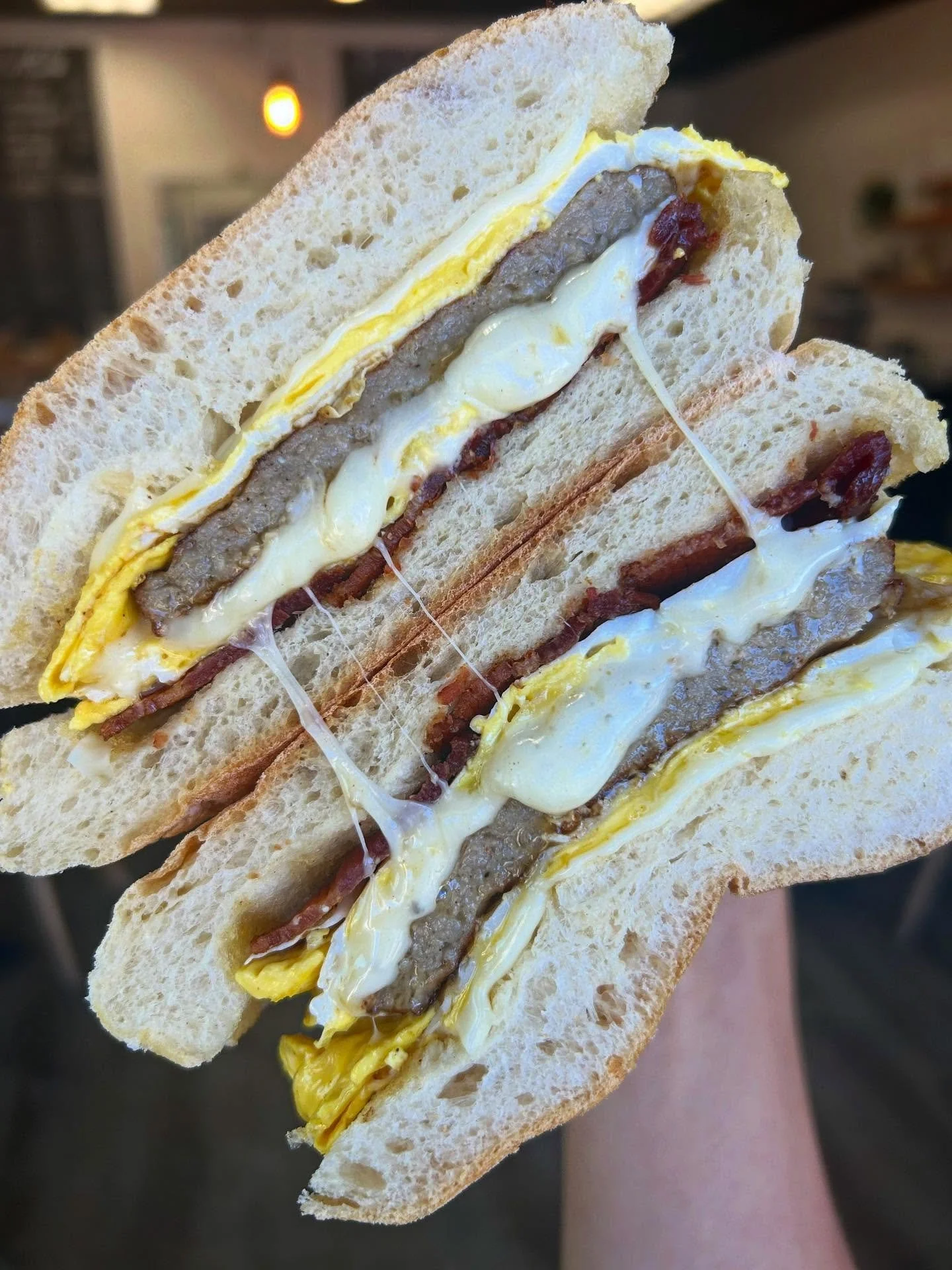 Close-up of a breakfast sandwich with egg, sausage, bacon, and melted cheese on white bread, held in hand.