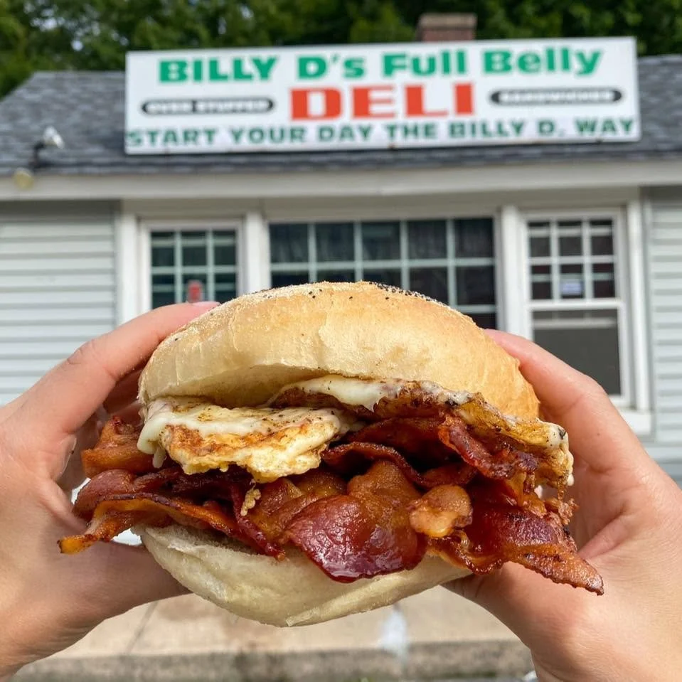 A hand holding a breakfast sandwich with bacon, egg, and cheese bun in front of a sign for Billy D's Full Belly Deli.