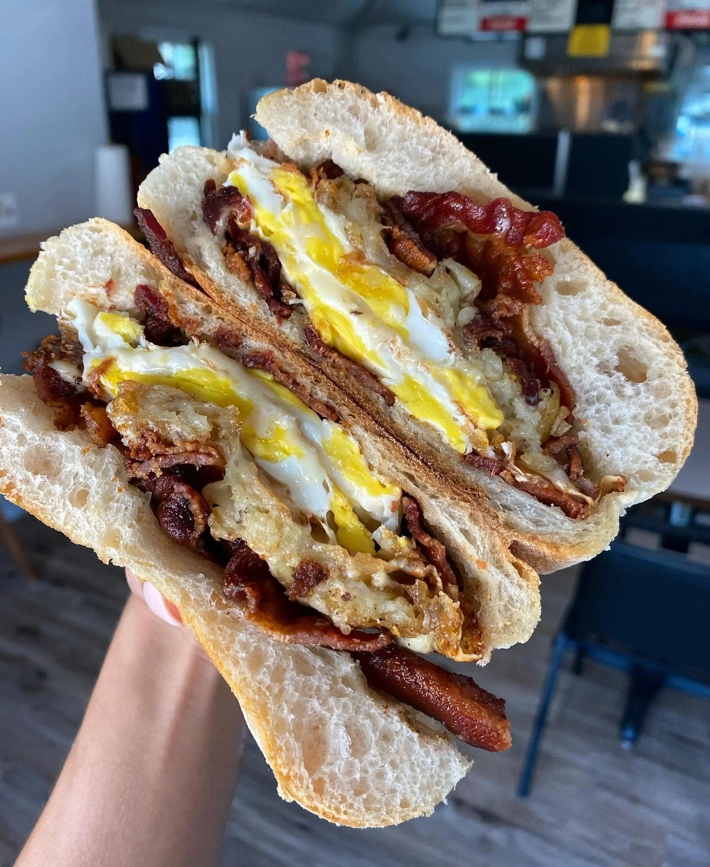 A sandwich with fried eggs, bacon, and possibly sausage or potato inside a baguette-style bread, held by a hand in a restaurant or café setting.