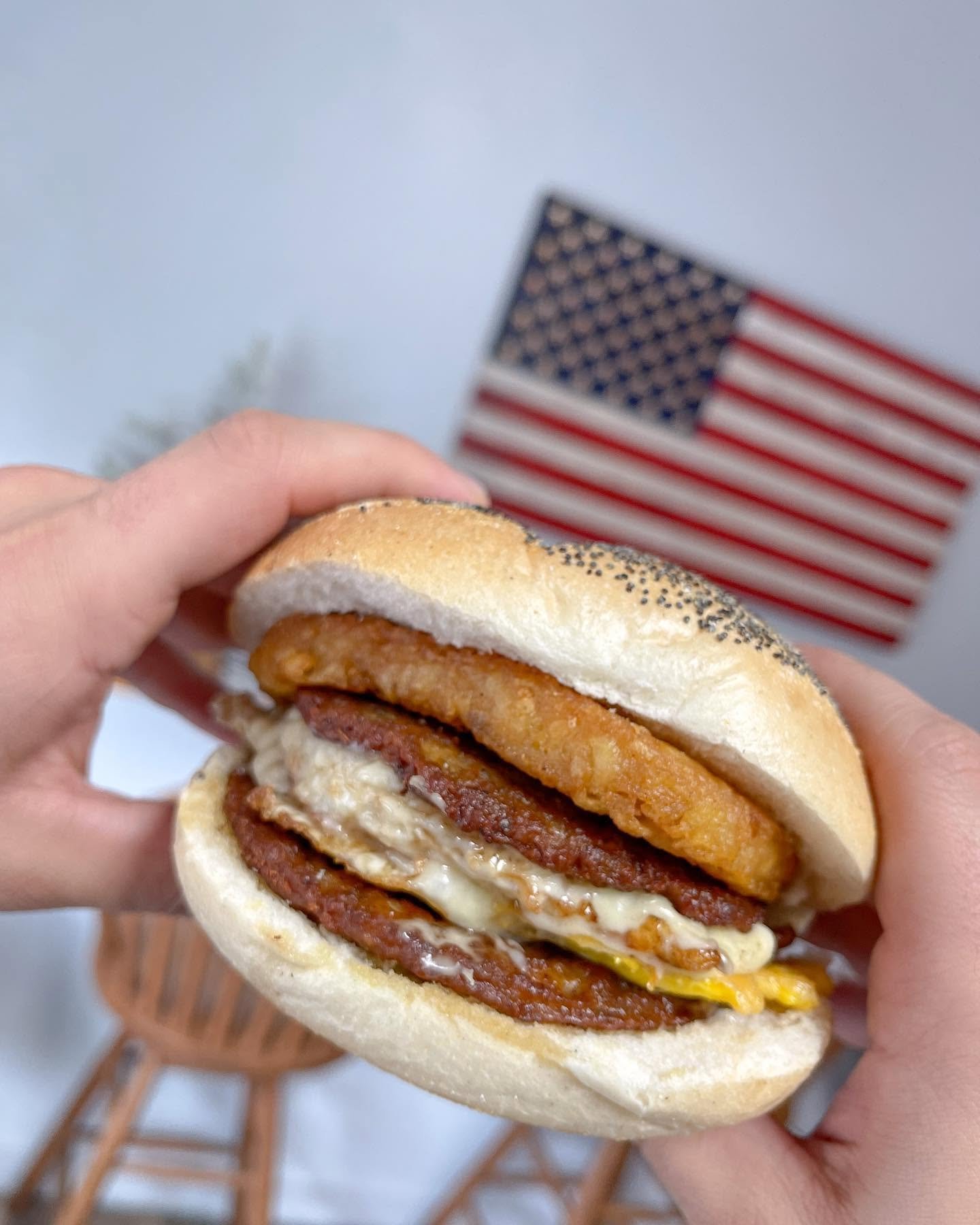 Hand holding a sandwich with fried chicken, bacon, melted cheese, and sauce in a sandwich bun, with an American flag in the background.