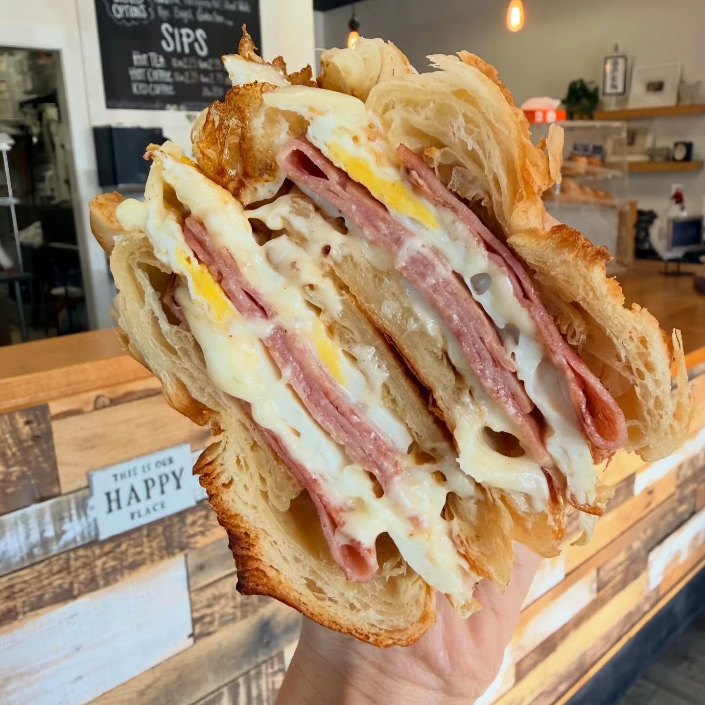 Close-up of a breakfast sandwich with layers of ham, egg, and cheese on toasted bread, held in hand inside a café.