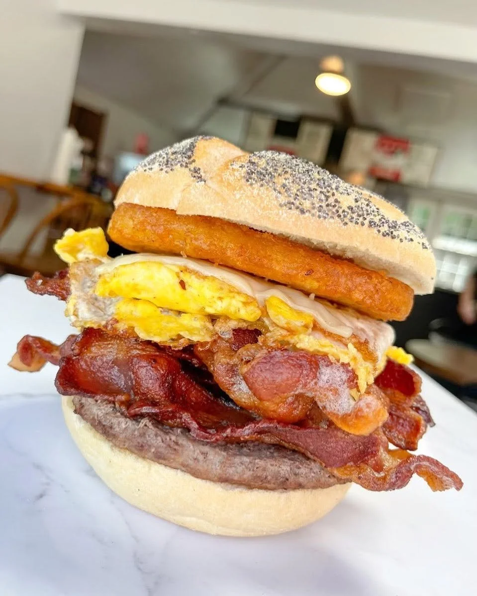 Close-up of a sandwich with bacon, eggs, hash brown, sausage, and poppy seed bun on a white surface in a dining area.