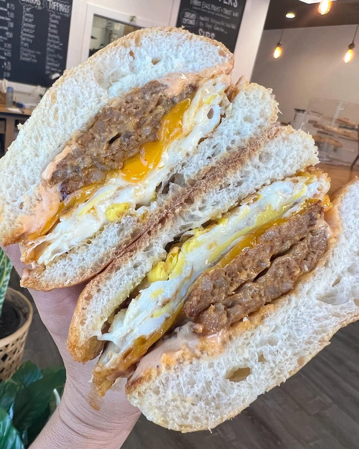 Close-up of a breakfast sandwich with fried eggs and sausage on toasted bread, held in a person's hand inside a cafe.