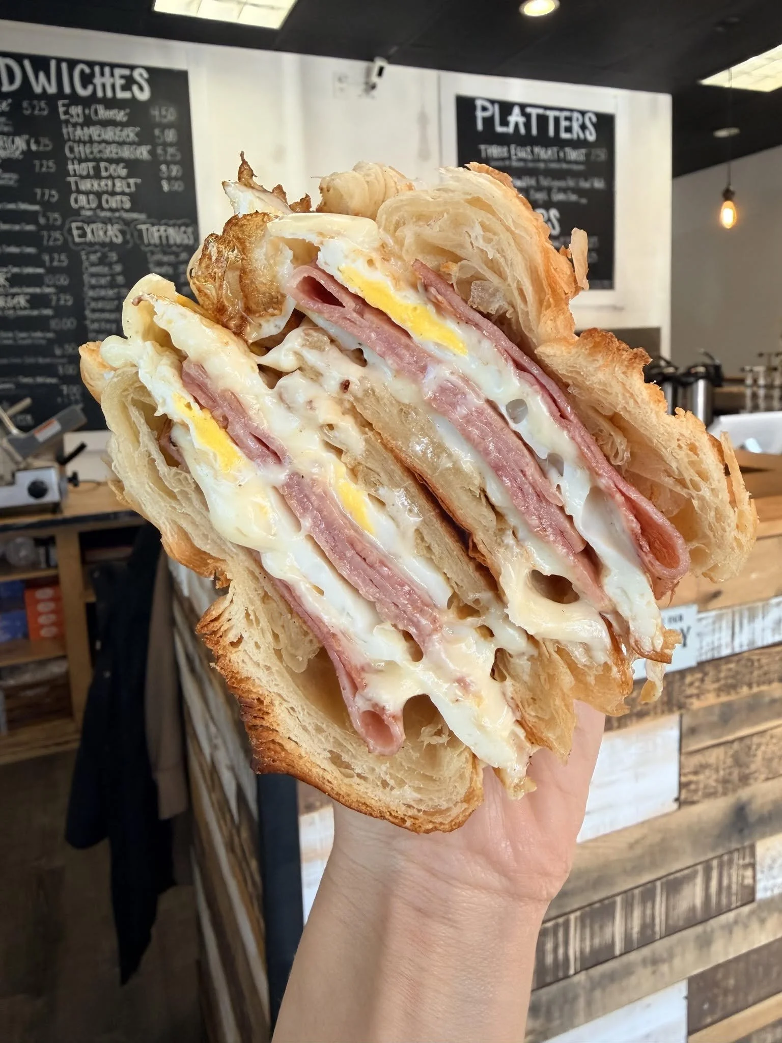 A person holding a sandwich with eggs, ham, and melted cheese in front of a menu board at a cafe.