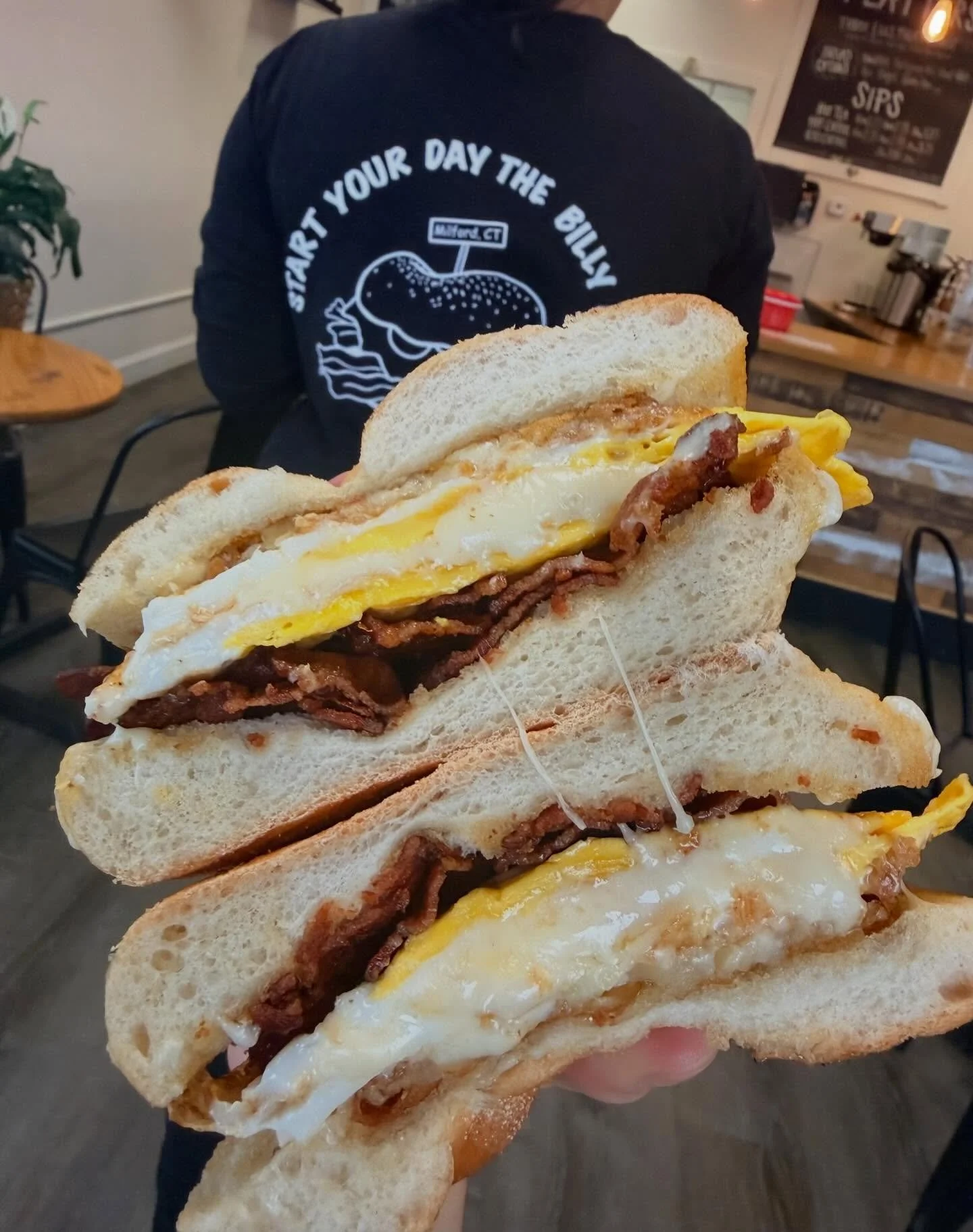 Close-up of a breakfast sandwich with eggs, bacon, with a person in the background wearing a black shirt with white text and a bun with sandwich fillings.