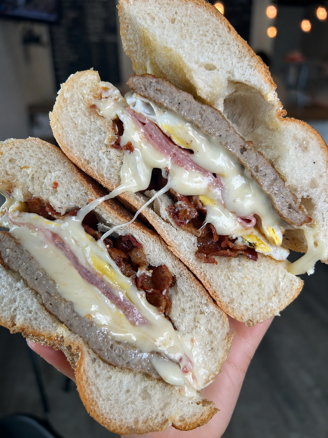 Close-up of a sandwich with layers of bacon, ham, cheese, and a sausage patty in a bread roll, with melted cheese.