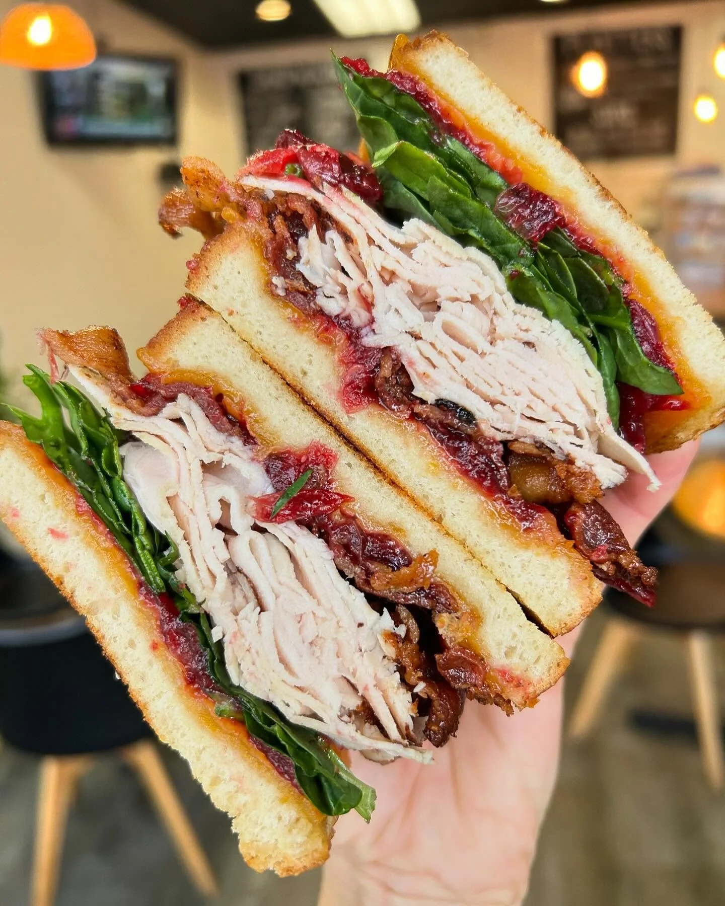 Close-up of a club sandwich with turkey, bacon, lettuce, tomato, and mayonnaise between toasted bread slices, held in hand in a restaurant or cafe setting.
