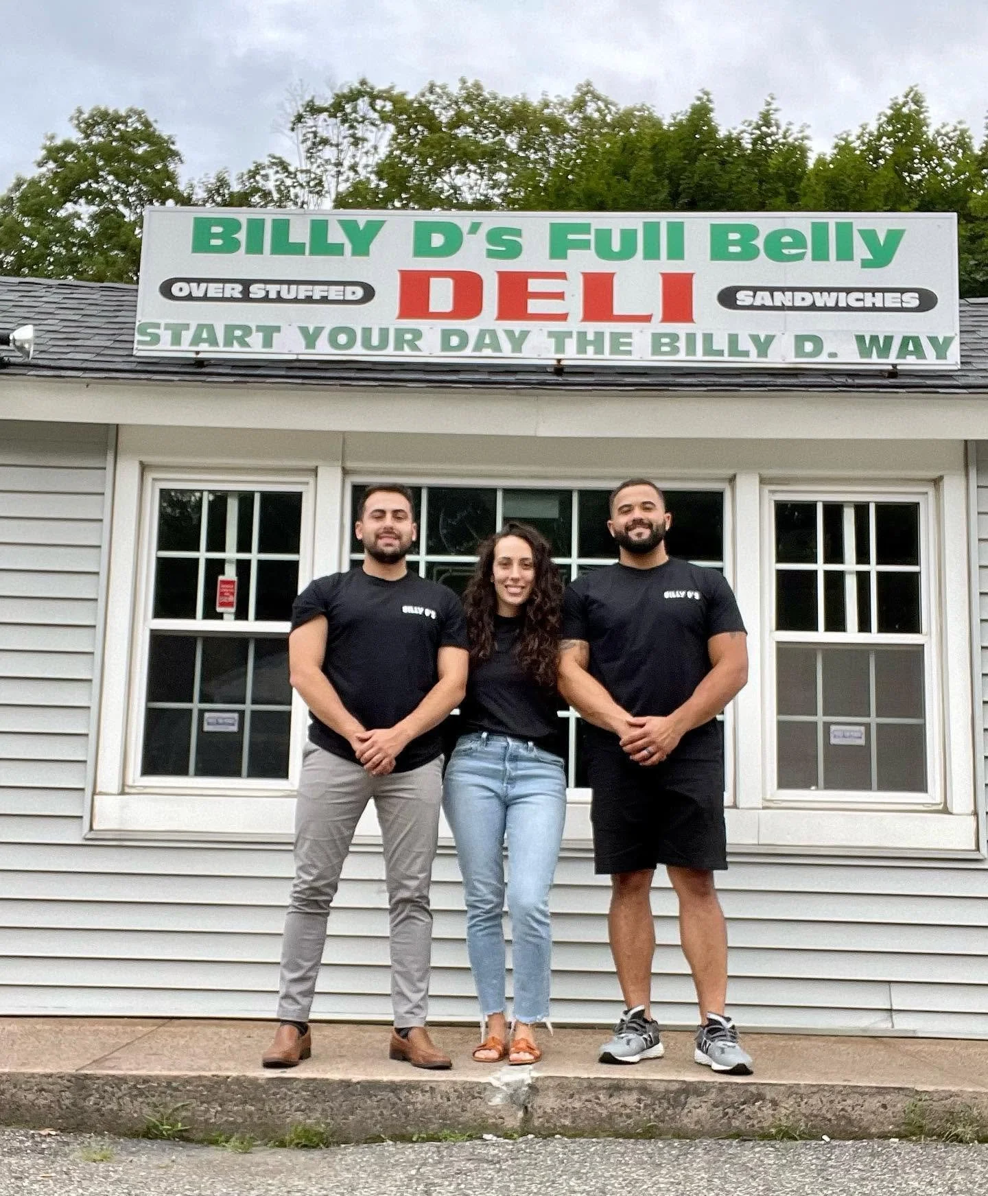 Three people standing in front of a building with a large sign that reads "Billy D's Full Belly Deli" and advertisements for sandwiches, overstuffed, start your day the Billy D. way.