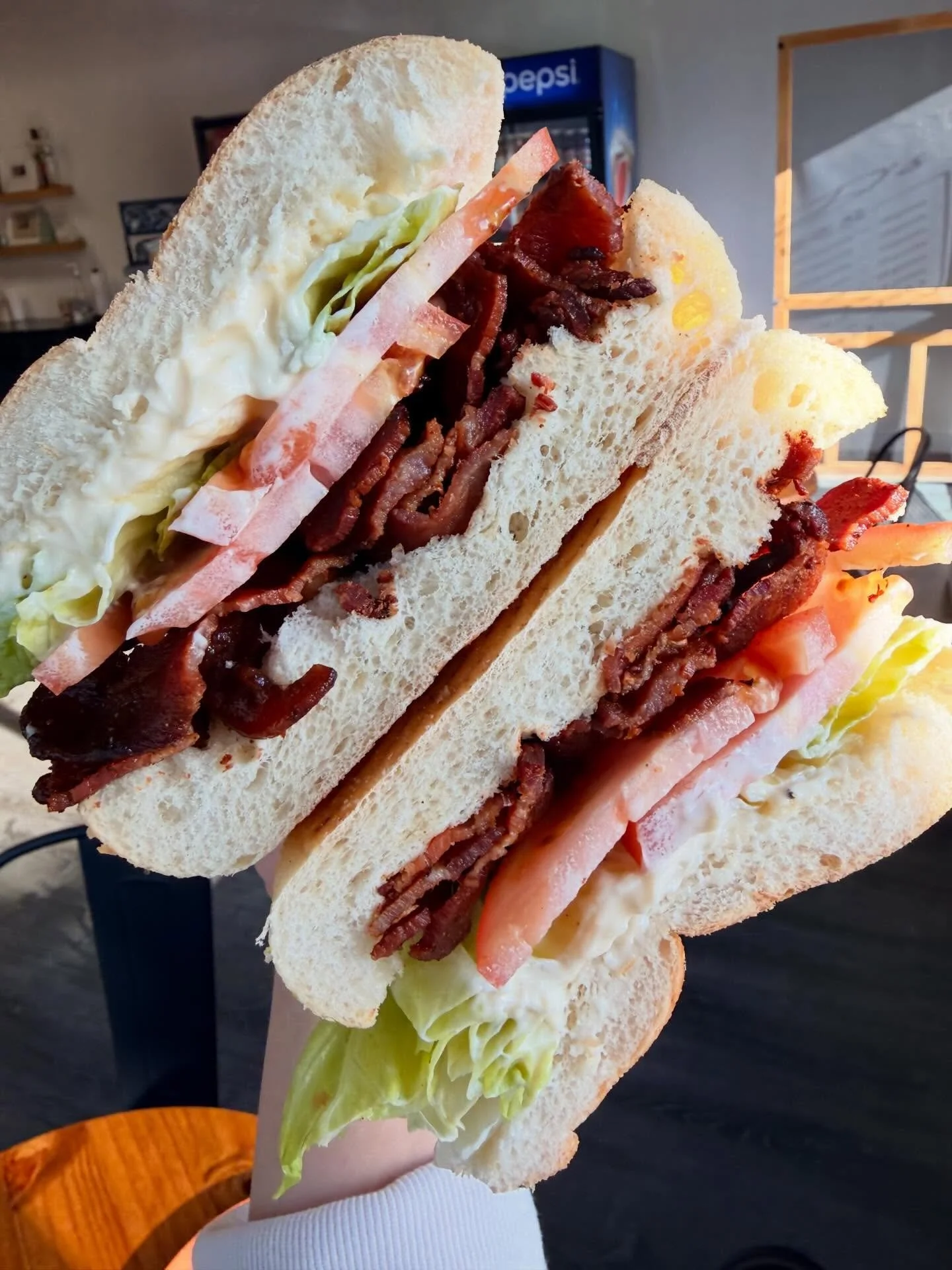 Close-up of a sandwich with lettuce, tomato, bacon, ham, and egg on white bread, held by a person in a restaurant setting.