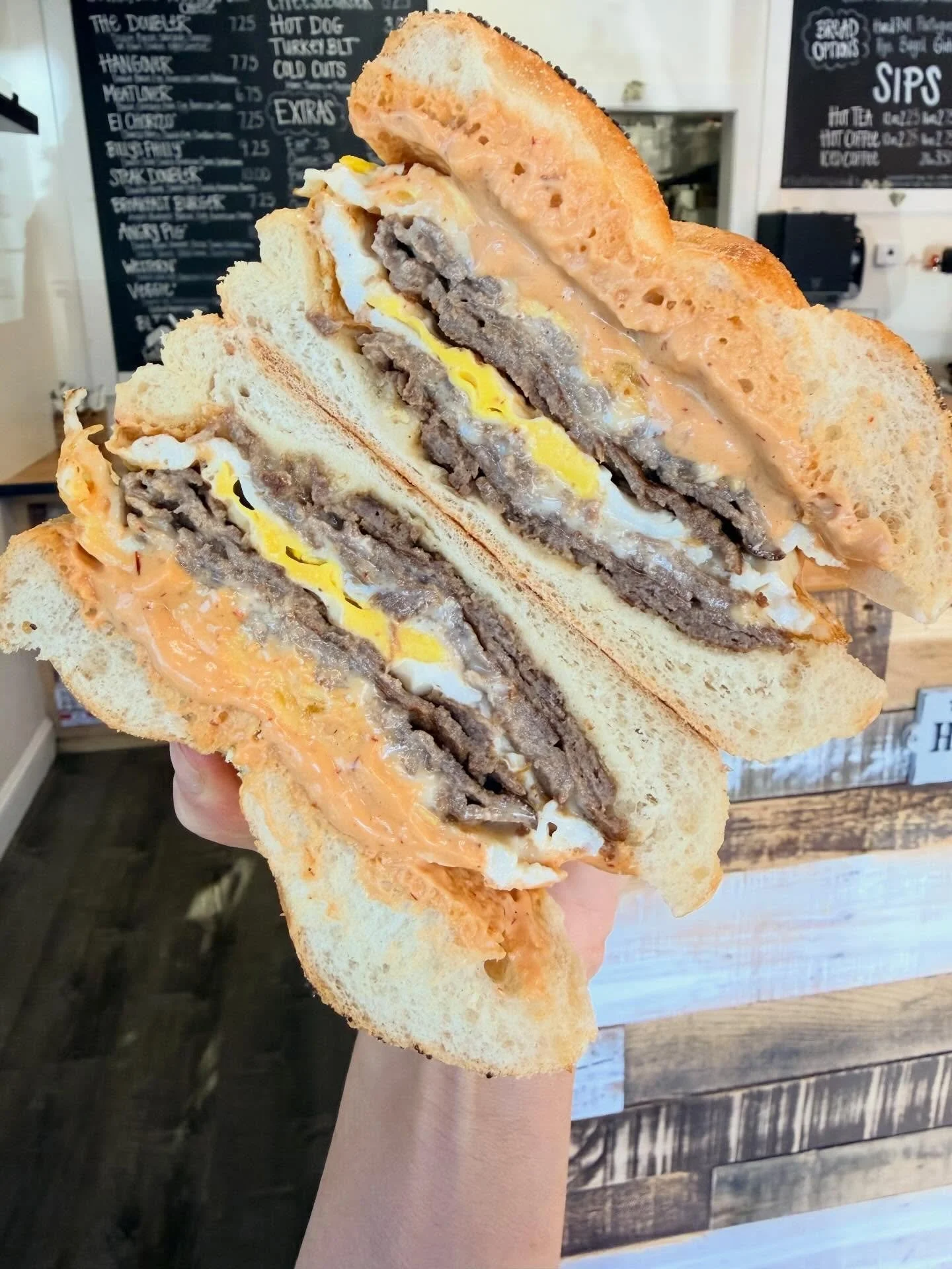 Two halves of a breakfast sandwich with sausage, egg, and cheese on toasted bread, held up in a person's hand inside a restaurant with a chalkboard menu in the background.
