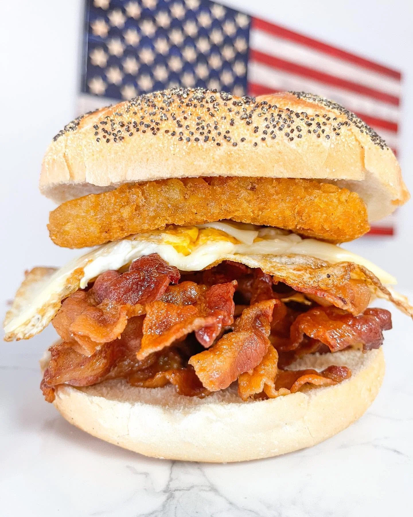 Close-up of a sandwich with bacon, fried egg, breaded fish fillet, and poppy seed bun, with an American flag in the background.