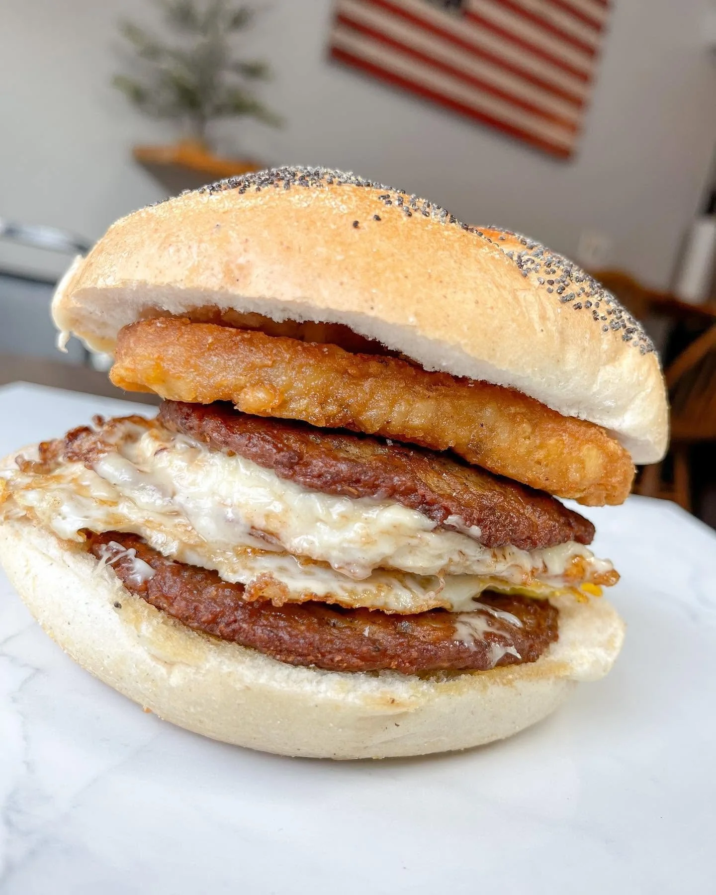 Close-up of a breakfast sandwich with fried chicken, bacon, and egg on an English muffin, with an American flag decoration in the background.