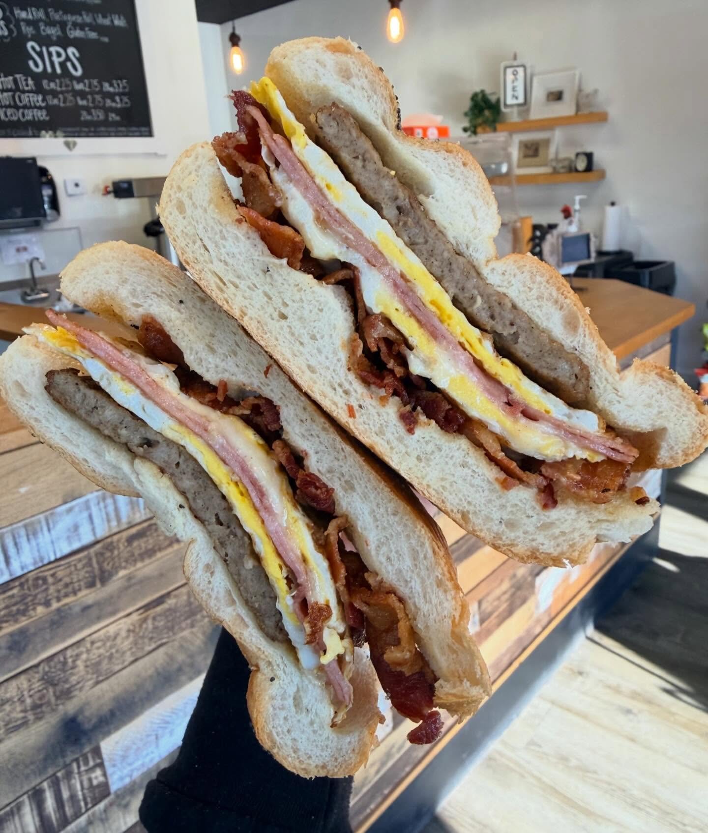 Stacked sandwiches with bacon, egg, ham, and bread inside a café with warm lighting and a chalkboard menu in the background.