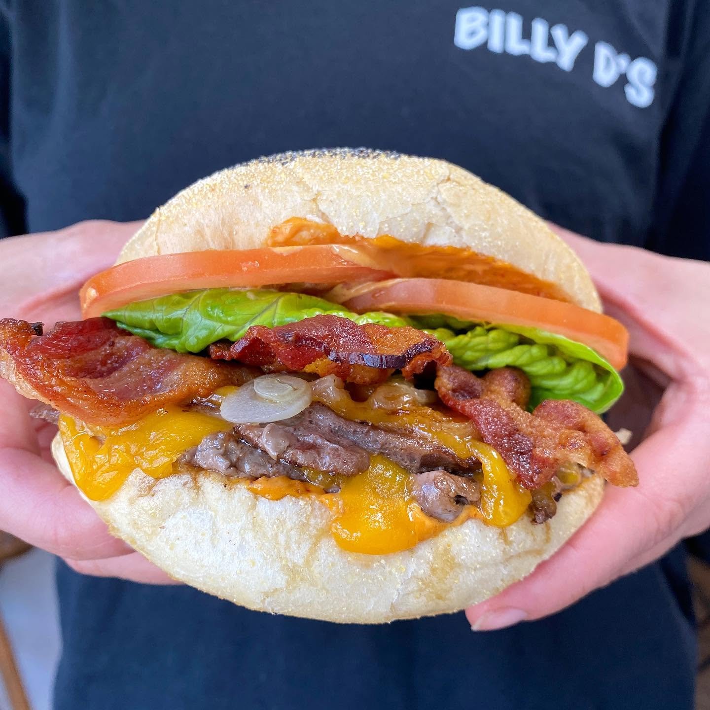 Close-up of a cheeseburger with bacon, lettuce, tomato, and onion held in a person's hand.