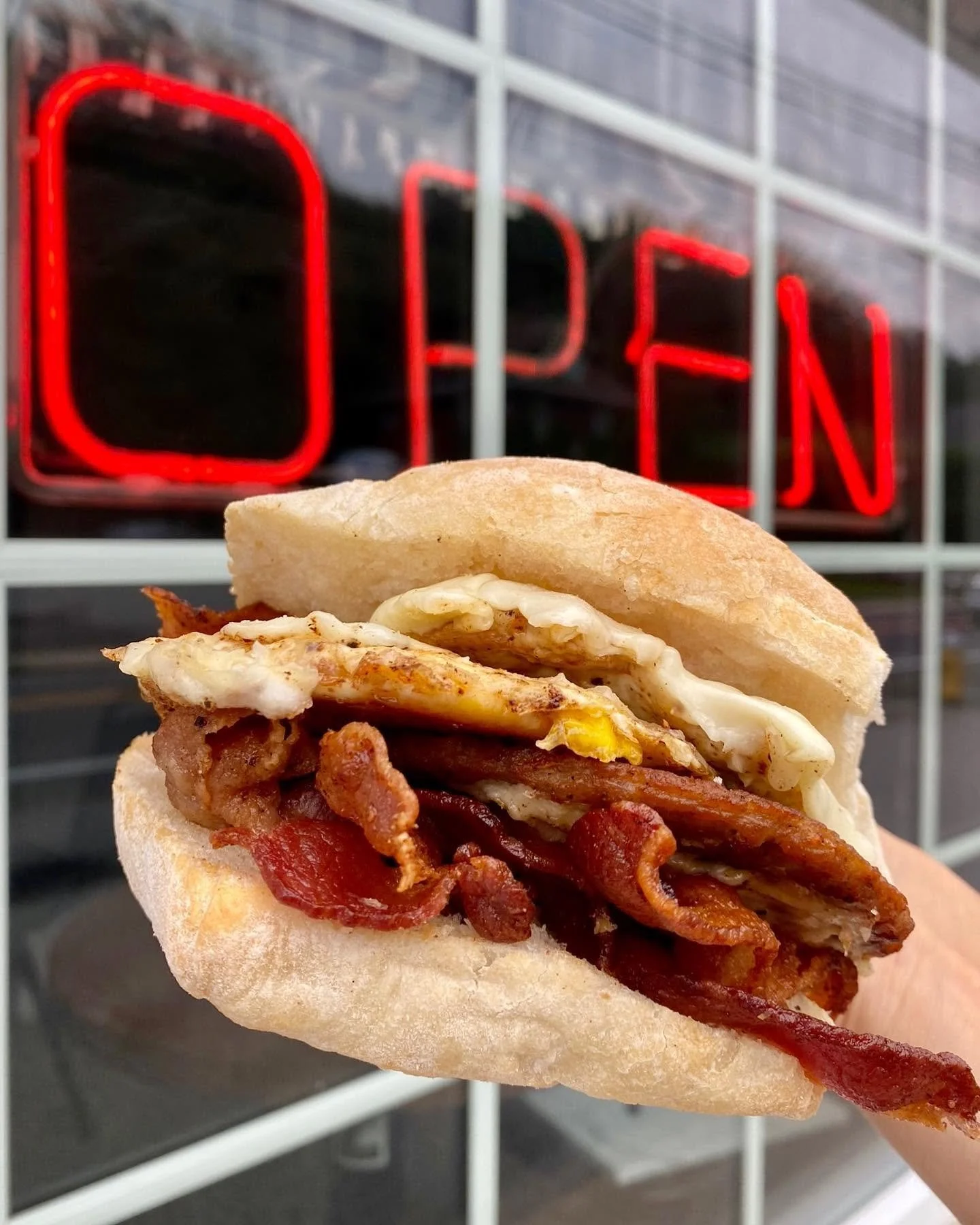 Bacon, egg, and bacon sandwich in a hoagie bun with a fast food restaurant "OPEN" sign in the background.