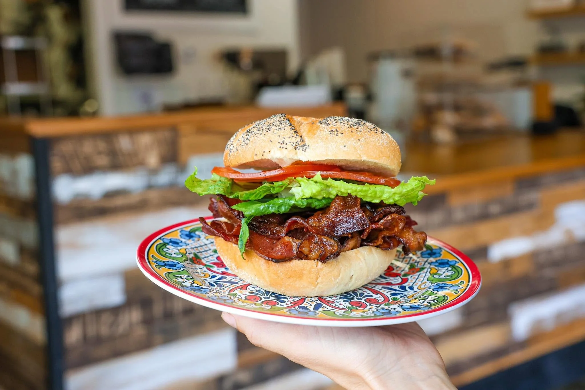 A plate holding a sandwich with crispy bacon, lettuce, tomato, and a bun with poppy seeds, held by a hand against a blurred wood and kitchen background.