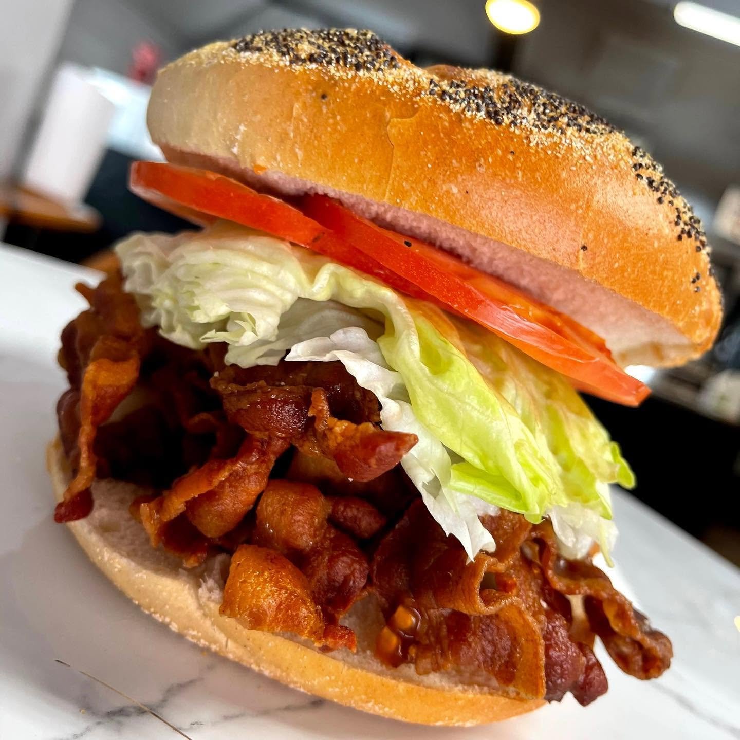 Close-up of a sandwich with bacon, lettuce, tomato, and a poppy seed bun, on a white plate.