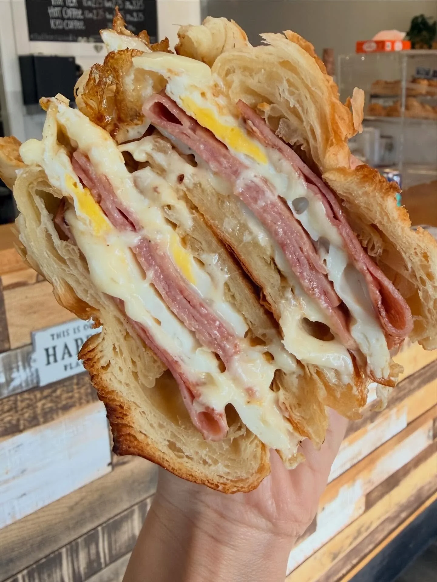 Close-up of a sandwich with eggs, ham, and cheese, held in a hand in front of a wooden display with baked goods.