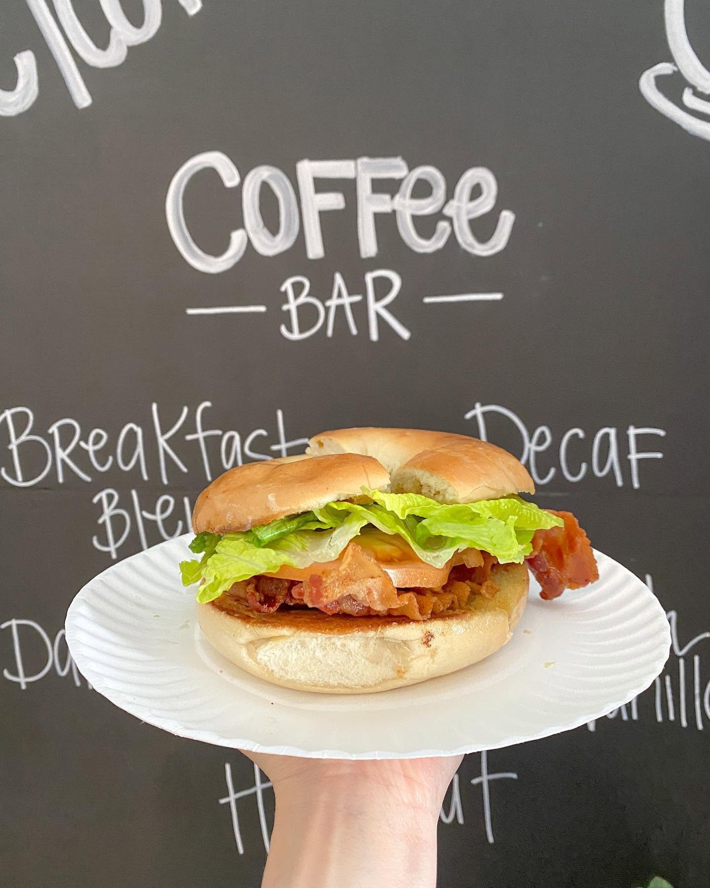 A person holding a paper plate with a breakfast sandwich in front of a chalkboard with 'Coffee Bar' written on it in white chalk.