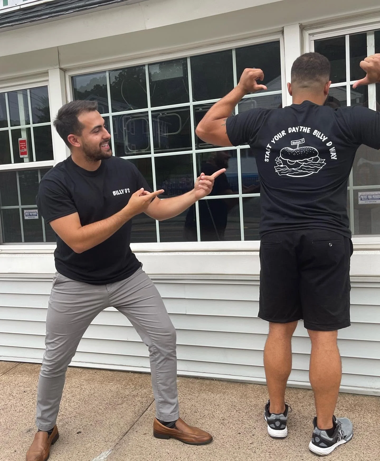 Two men standing outside in front of a house with large windows, posing as if flexing their arm muscles. One man is smiling and pointing at the other.