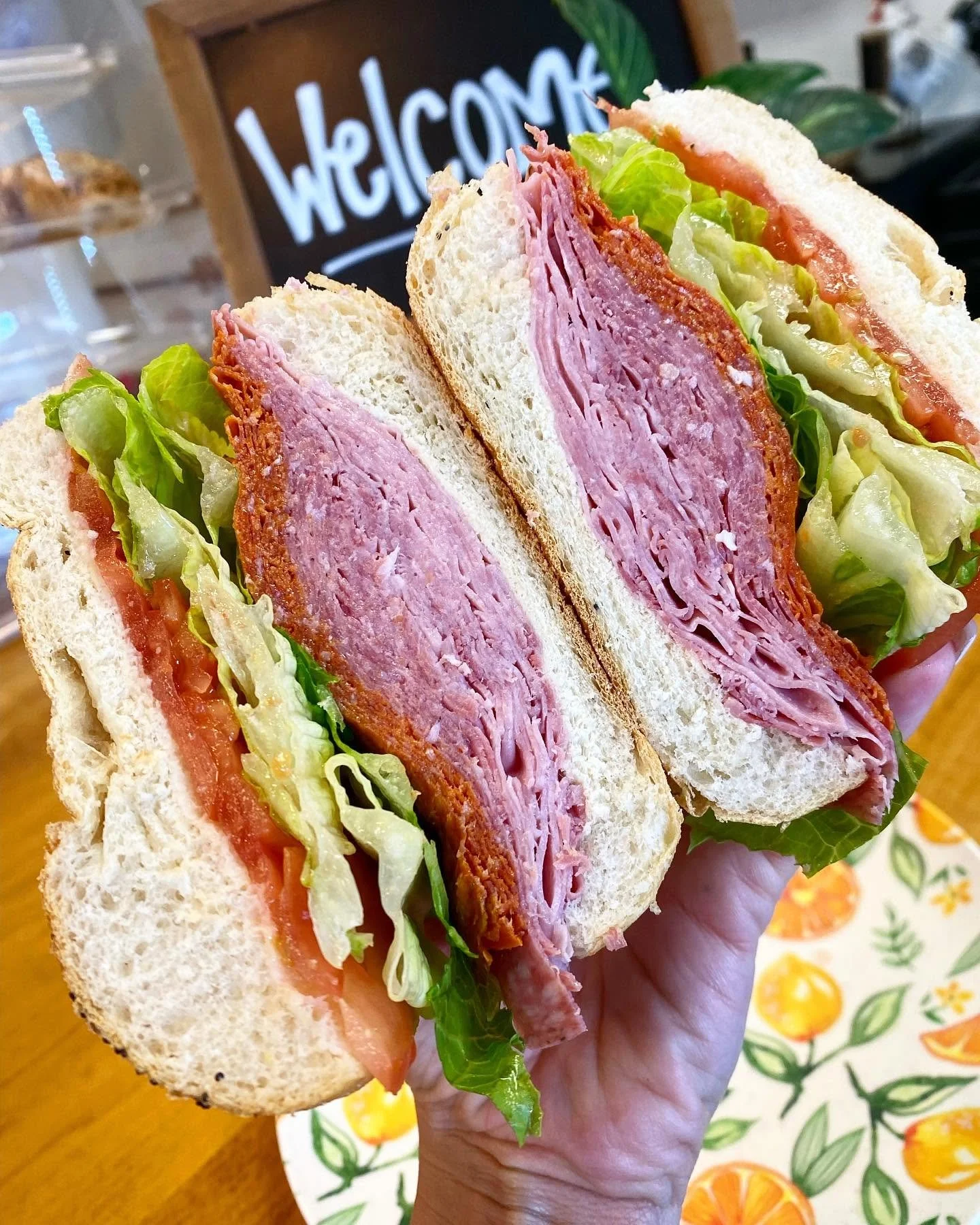 Close-up of a hand holding a sandwich with lettuce, tomato, slices of ham, and bread. In the background, a blackboard with the word 'Welcome' written in white.