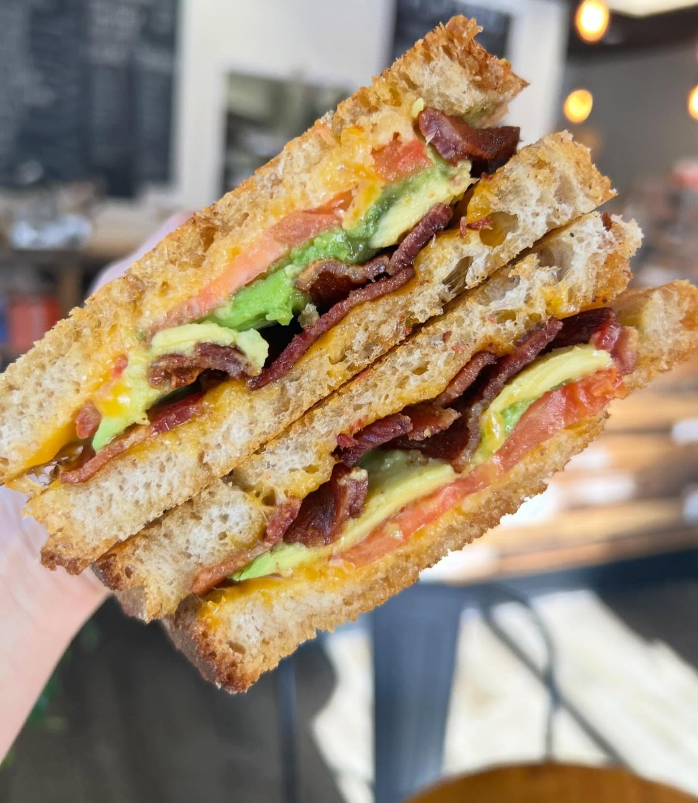 Close-up of a sandwich with toasted bread, bacon, lettuce, tomato, cheese, and egg, held in a hand with a blurred restaurant background.