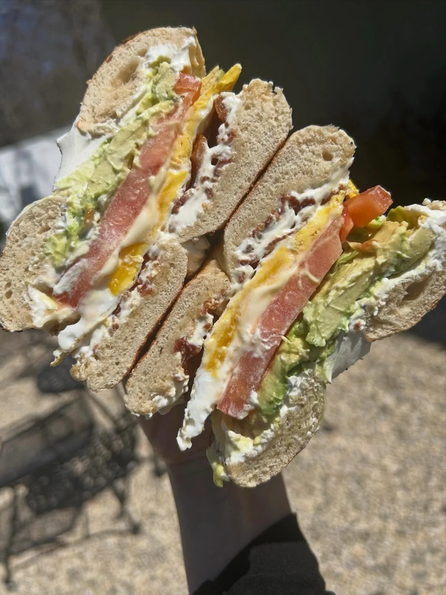 A sandwich with lettuce, tomato, egg, bacon, and mayonnaise on whole wheat bread, held in a person's hand outdoors.