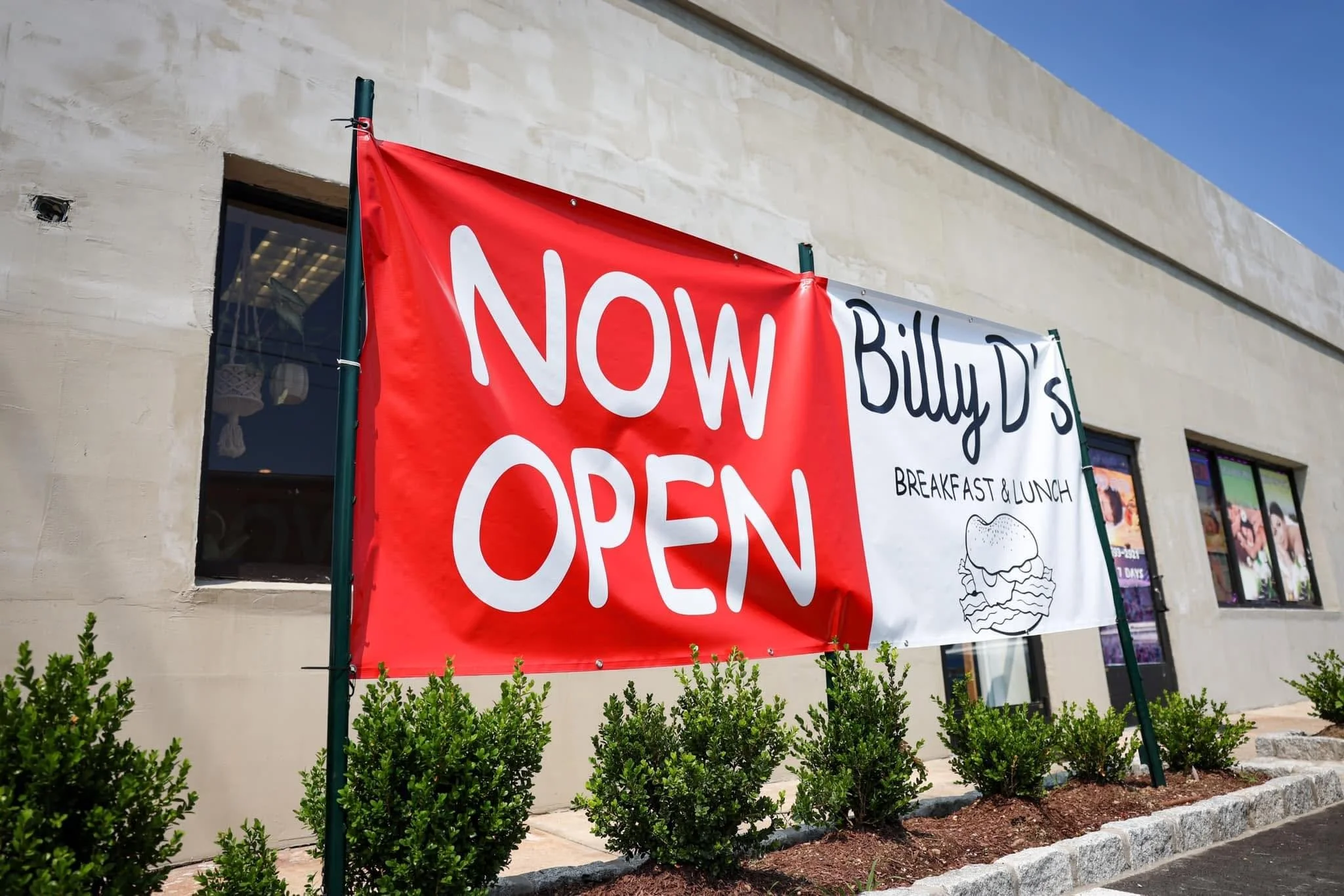 A building with a large red and white banner that reads 'Now Open' for a breakfast and lunch restaurant. The banner has a small drawing of a sandwich. There are green bushes and a sidewalk in front of the building.