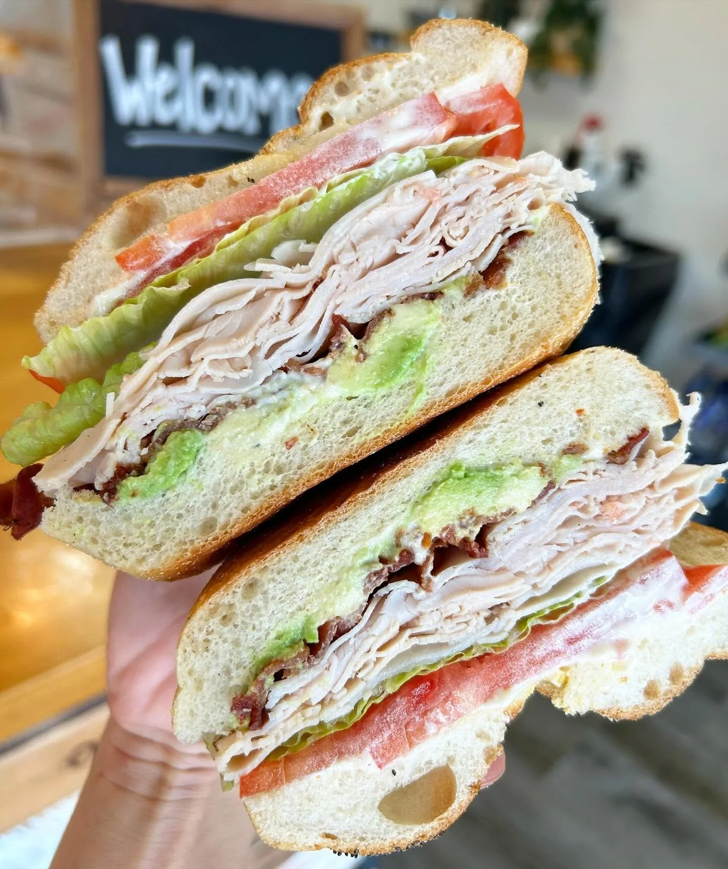 A close-up of a sliced turkey sandwich with lettuce, tomato, bacon, and mayonnaise on toasted white bread, being held in a person's hand.