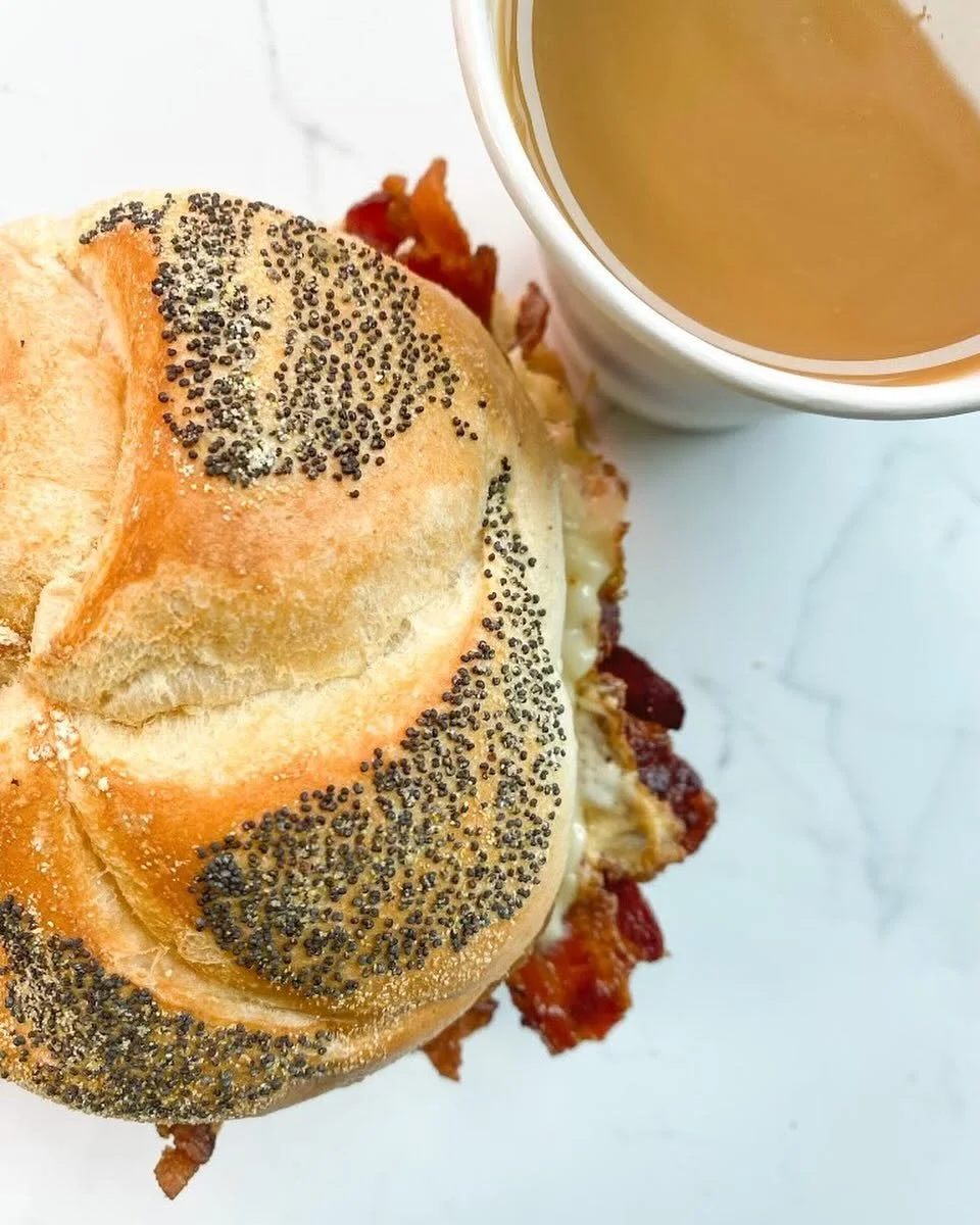 Close-up of a breakfast sandwich with poppy seed bun, bacon, cheese, and egg, next to a cup of coffee on a white marble surface.