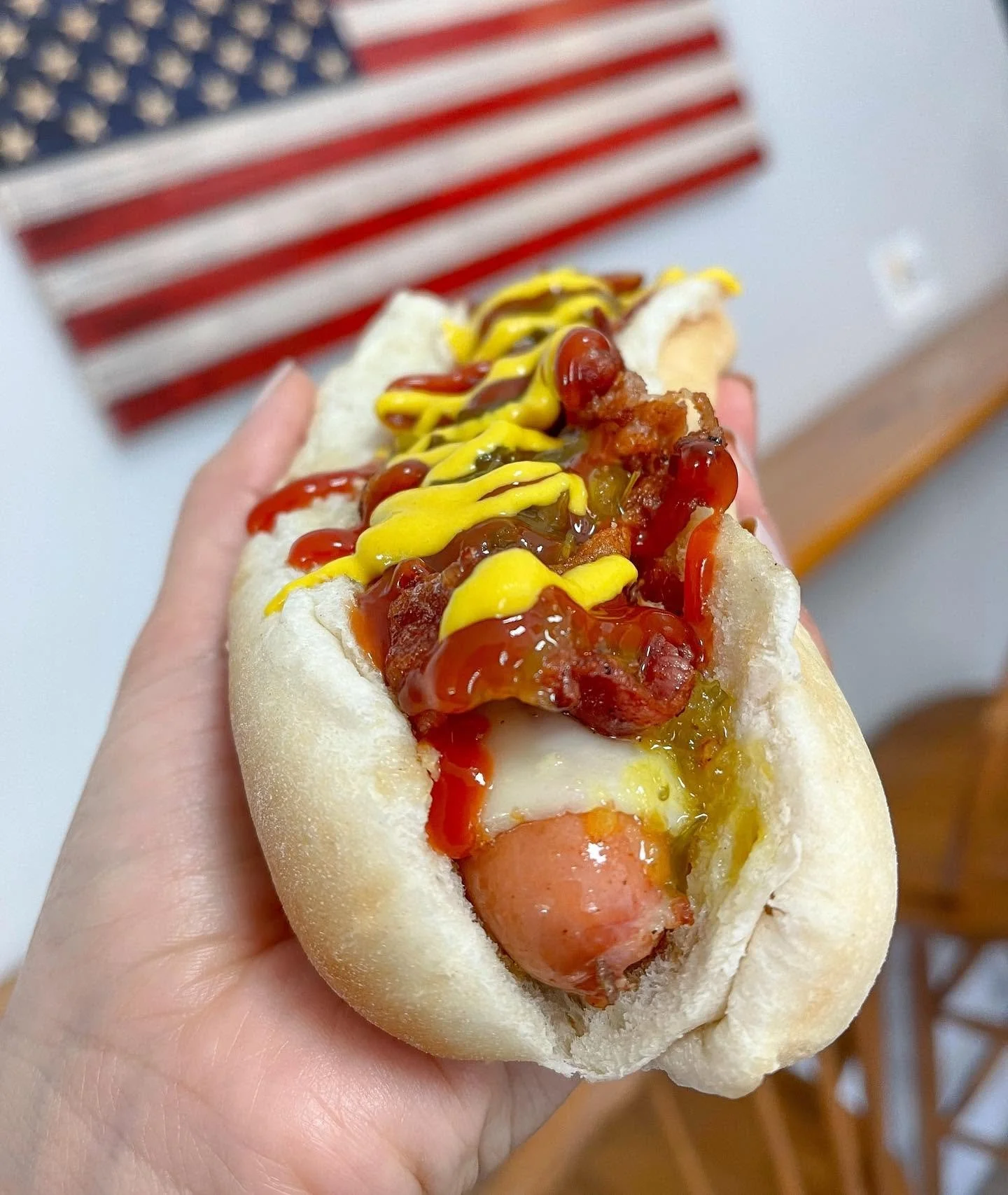 Close-up of a hot dog topped with ketchup, mustard, and relish, held in a person's hand. In the background, part of a U.S. flag is visible.