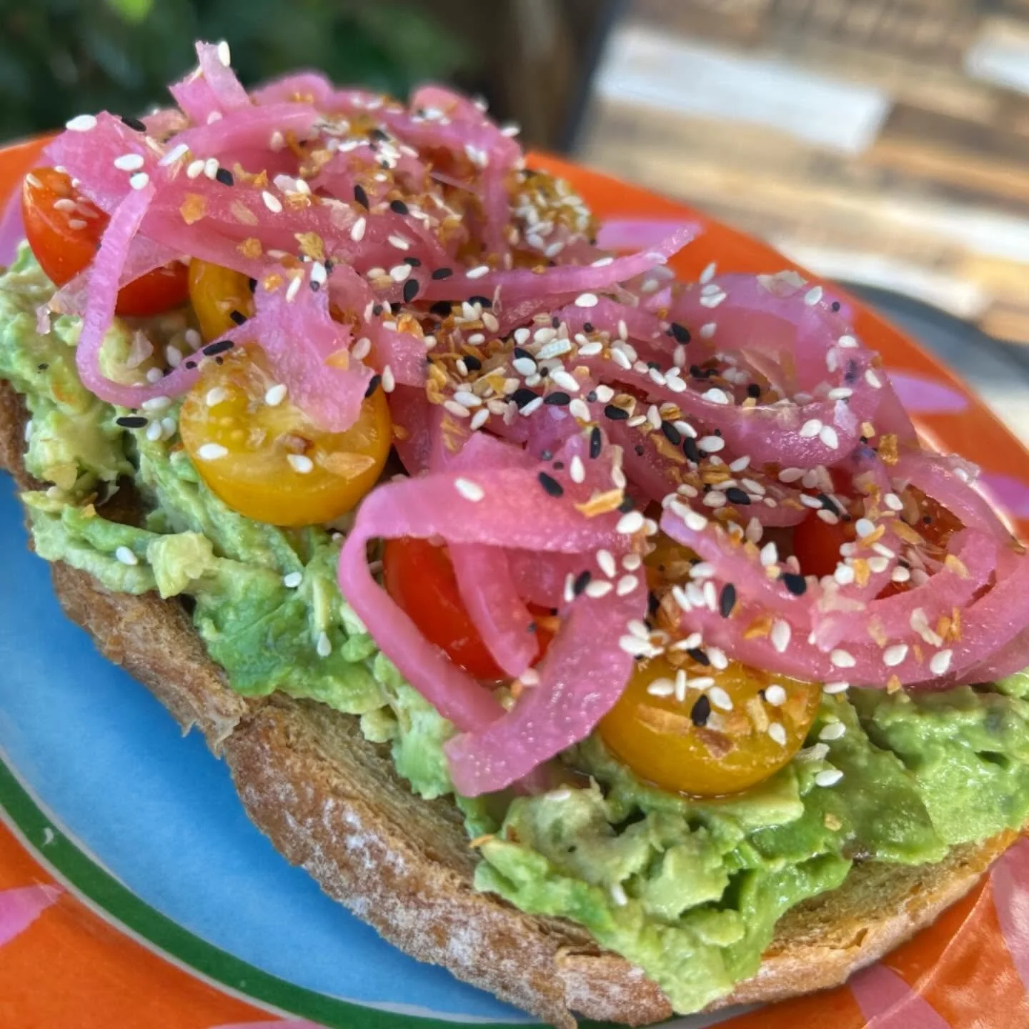 Close-up of a slice of avocado toast topped with cherry tomatoes, pickled red onions, and sesame seeds.