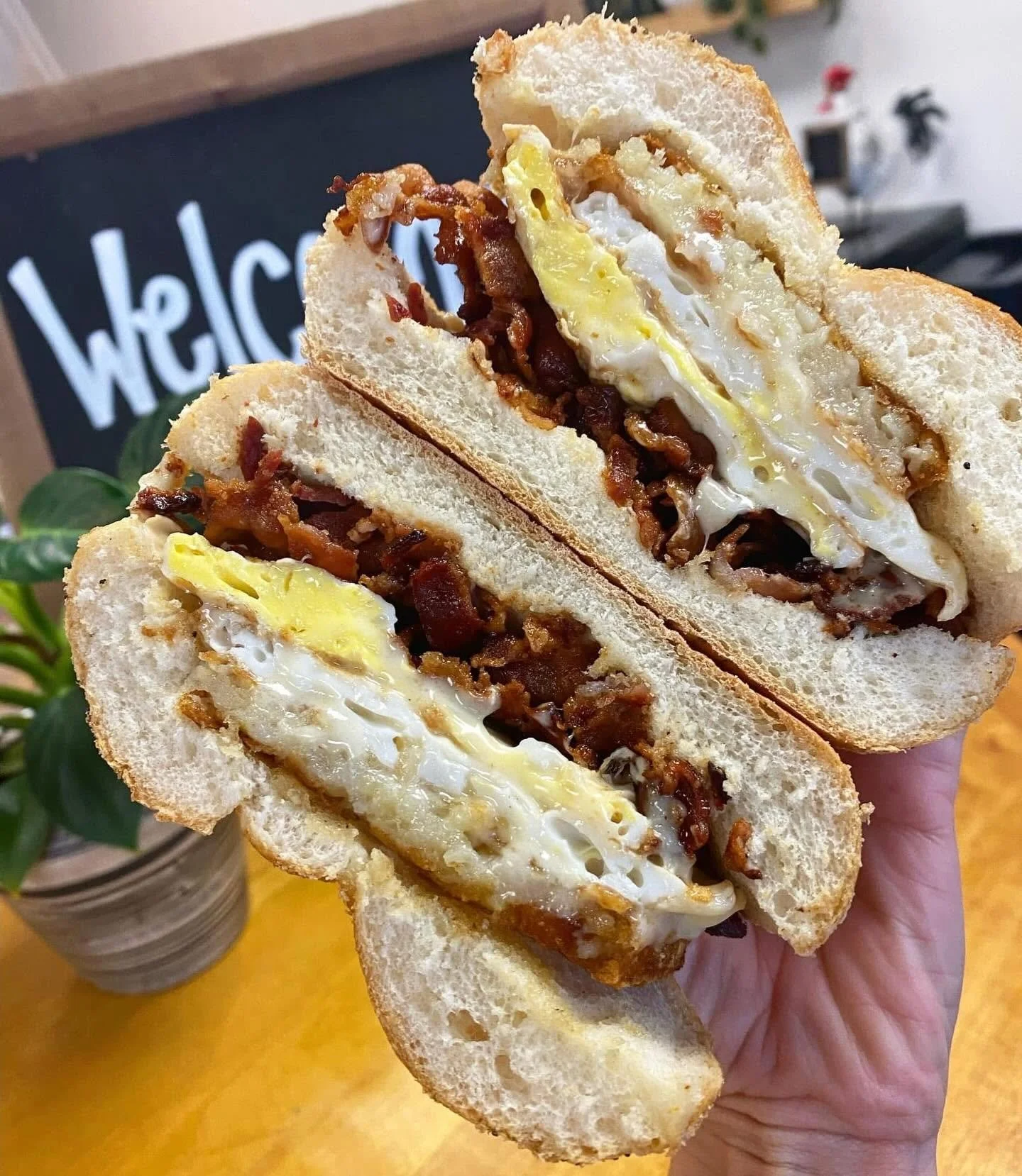 A hand holding a breakfast sandwich with fried egg, bacon, and meat filling on toasted bread.
