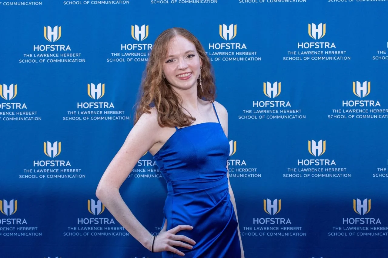A young woman with red hair in a blue dress smiling in front of a blue backdrop with the logo and name of Hofstra University, the Lawrence Herbert School of Communication.