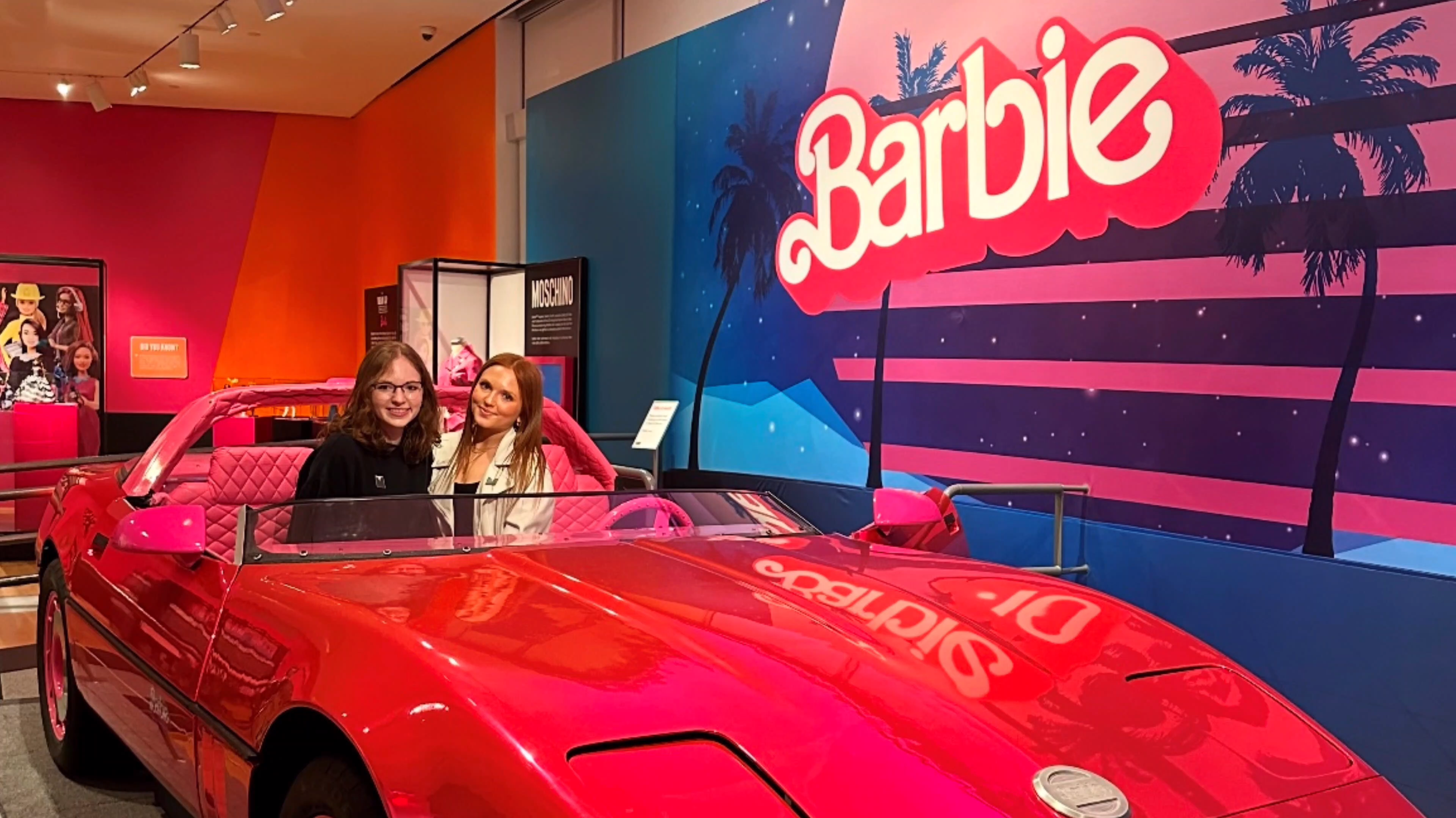 Two young women sitting in a bright pink toy car in front of a colorful display with palm trees and the word 'Barbie' on a sign, at an indoor exhibit or store.