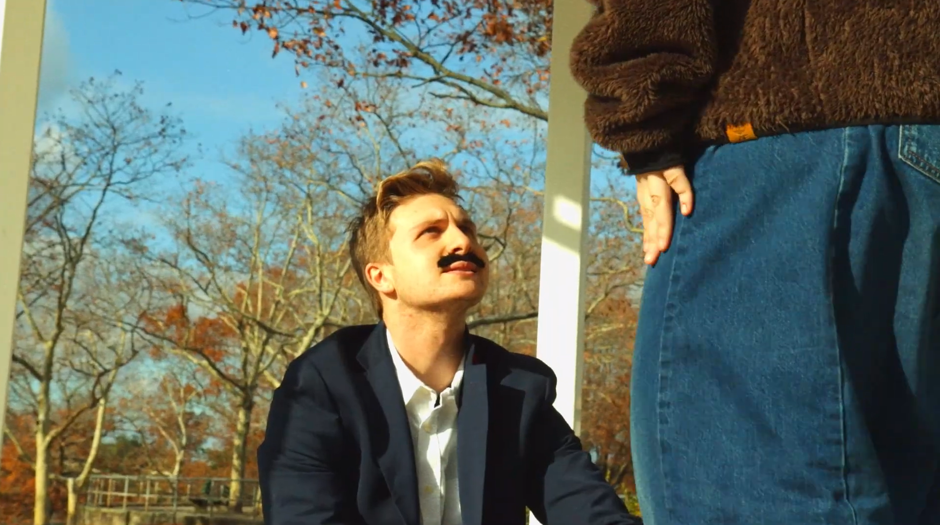 A man with a mustache wearing a suit and white shirt, looking up at another person outside during autumn. The other person is wearing blue jeans and a brown sweater, with their hand resting on their knee. The background shows leafless trees against a clear sky.
