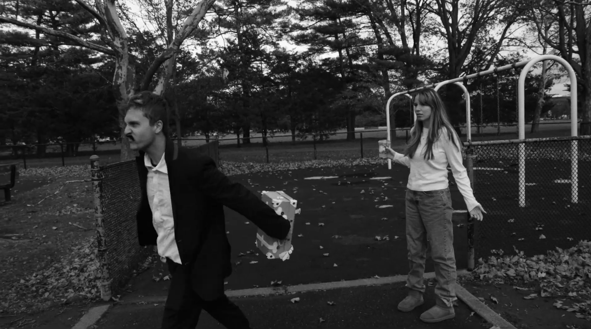 A black-and-white photo of a park with two young people, a man and a woman. The man, on the left, is wearing a suit and walking quickly, with a serious or angry expression, holding a stack of blocks. The woman, on the right, stands calmly near a playground structure, holding a drink, with a neutral expression. The scene is set among trees and fallen leaves.