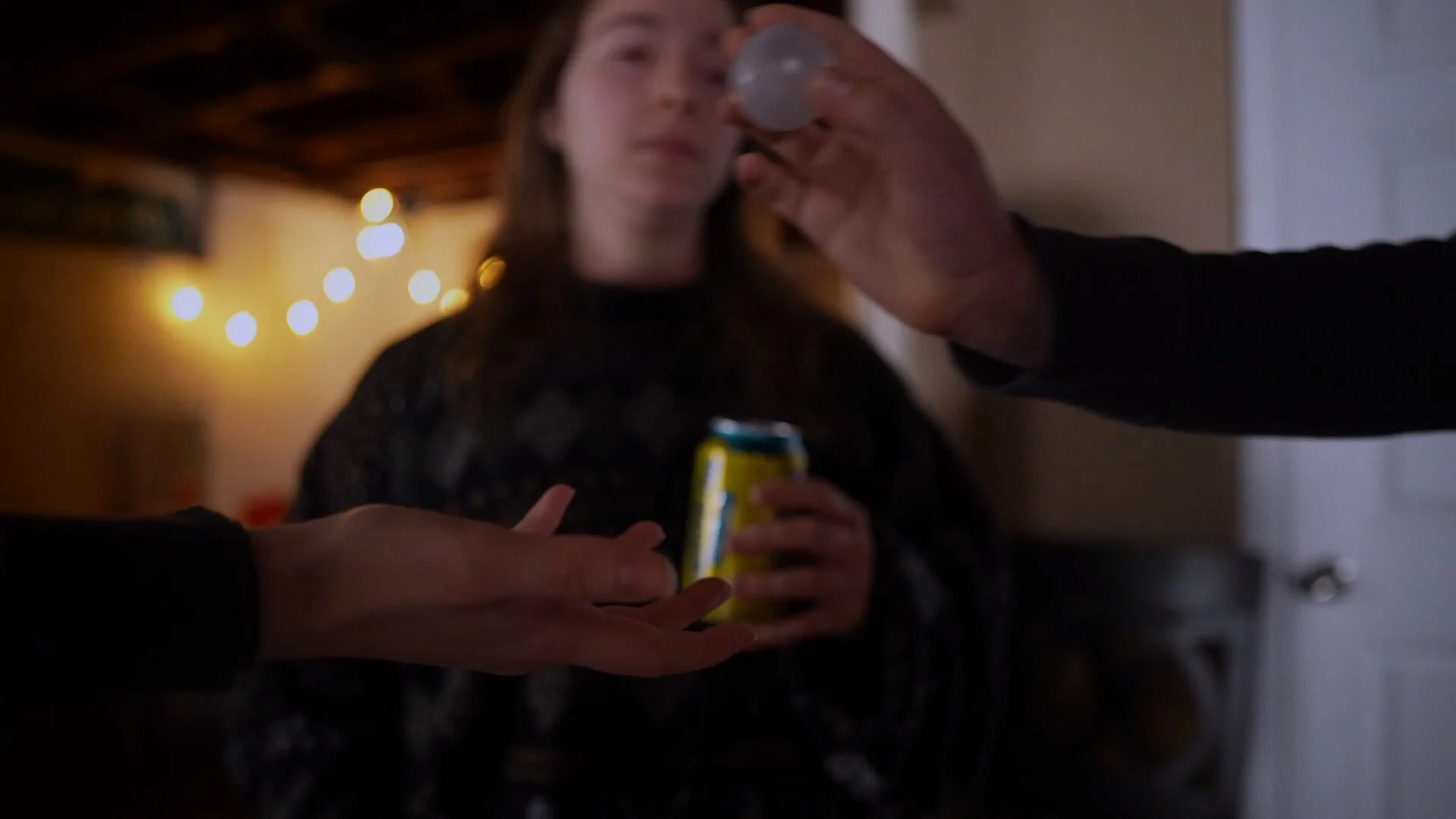 A woman holding a soda can with a person pouring a drink into her mouth at a dimly lit gathering.