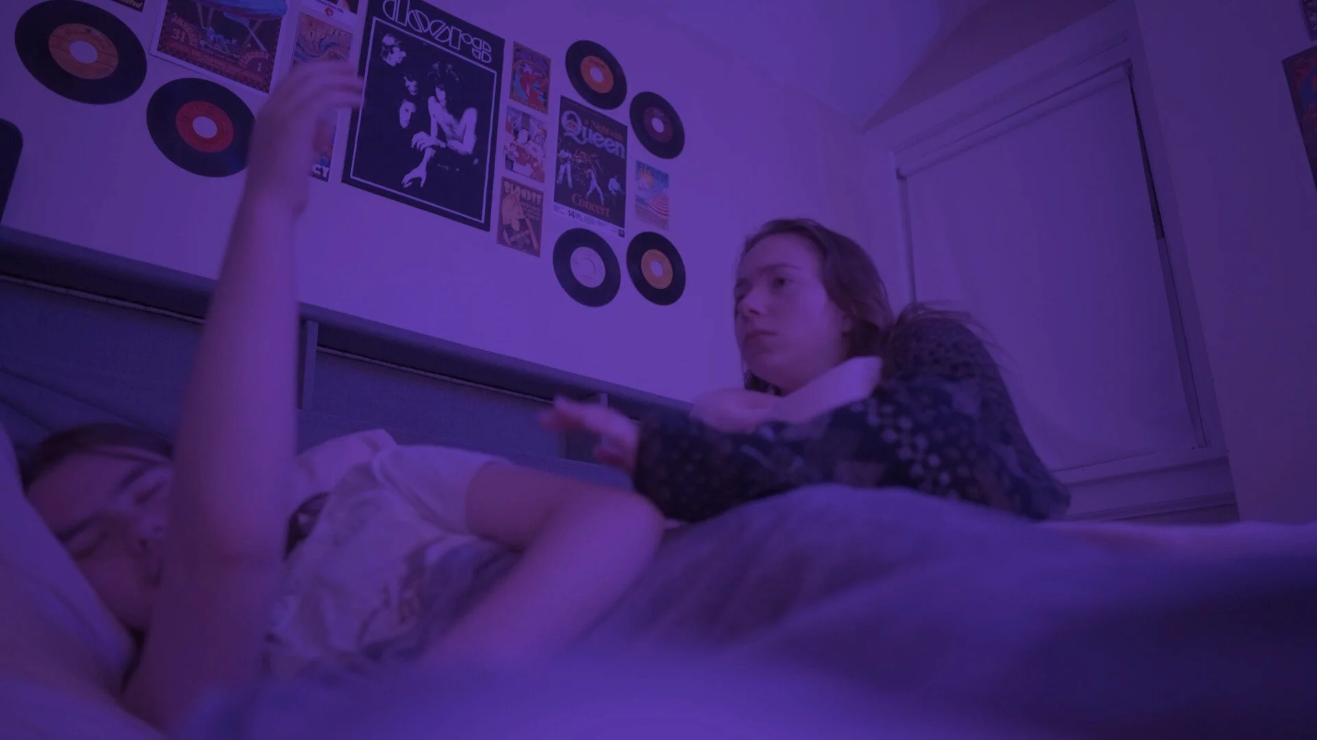 A girl lying on a bed with a purple lighting, looking at a person laying beside her, with a wall decorated with vinyl records and posters in the background.