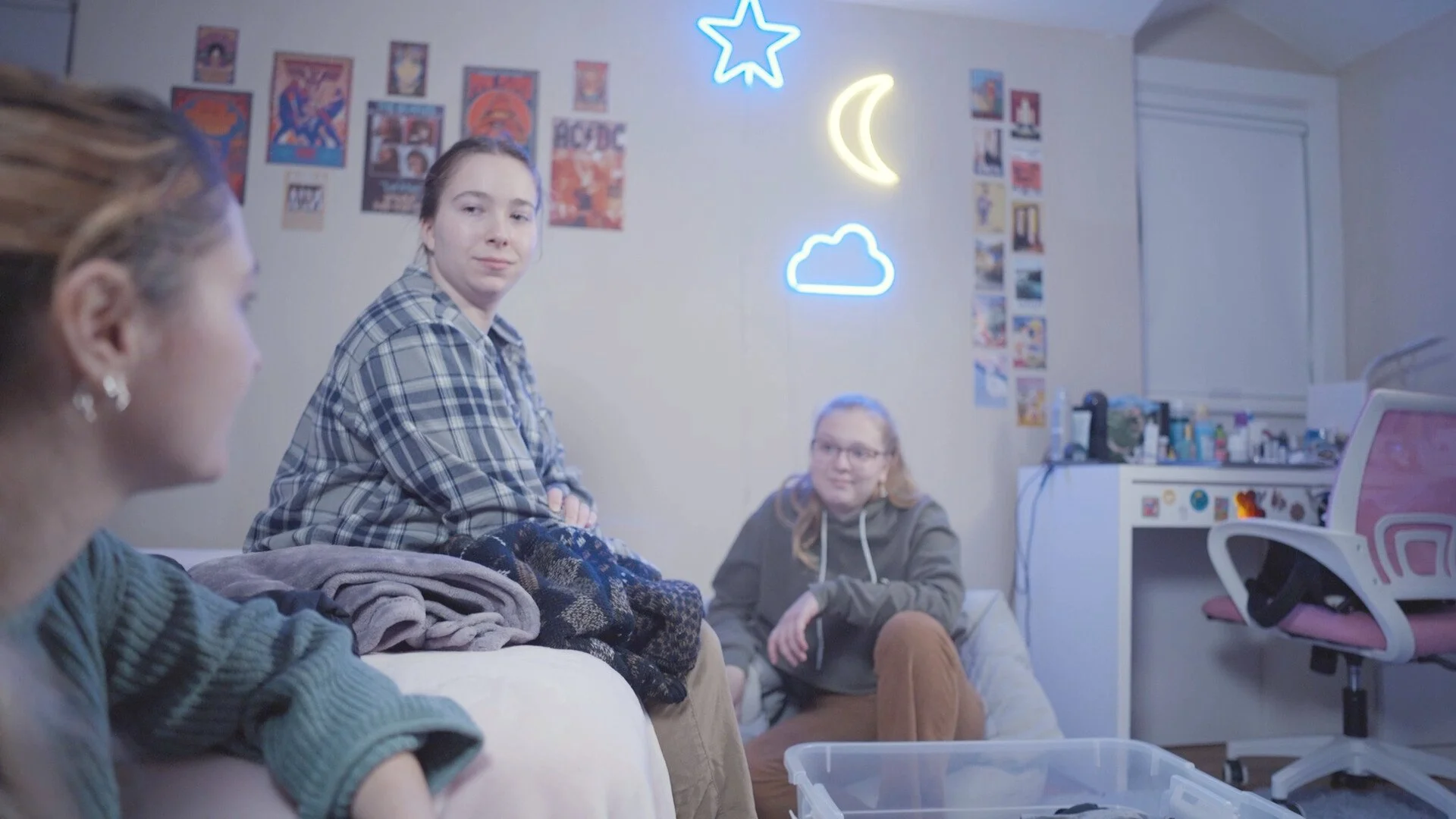 Three young women in a bedroom engaged in conversation, with neon cloud, star, moon, and cloud signs on the wall behind them, and various posters and photos on the wall.