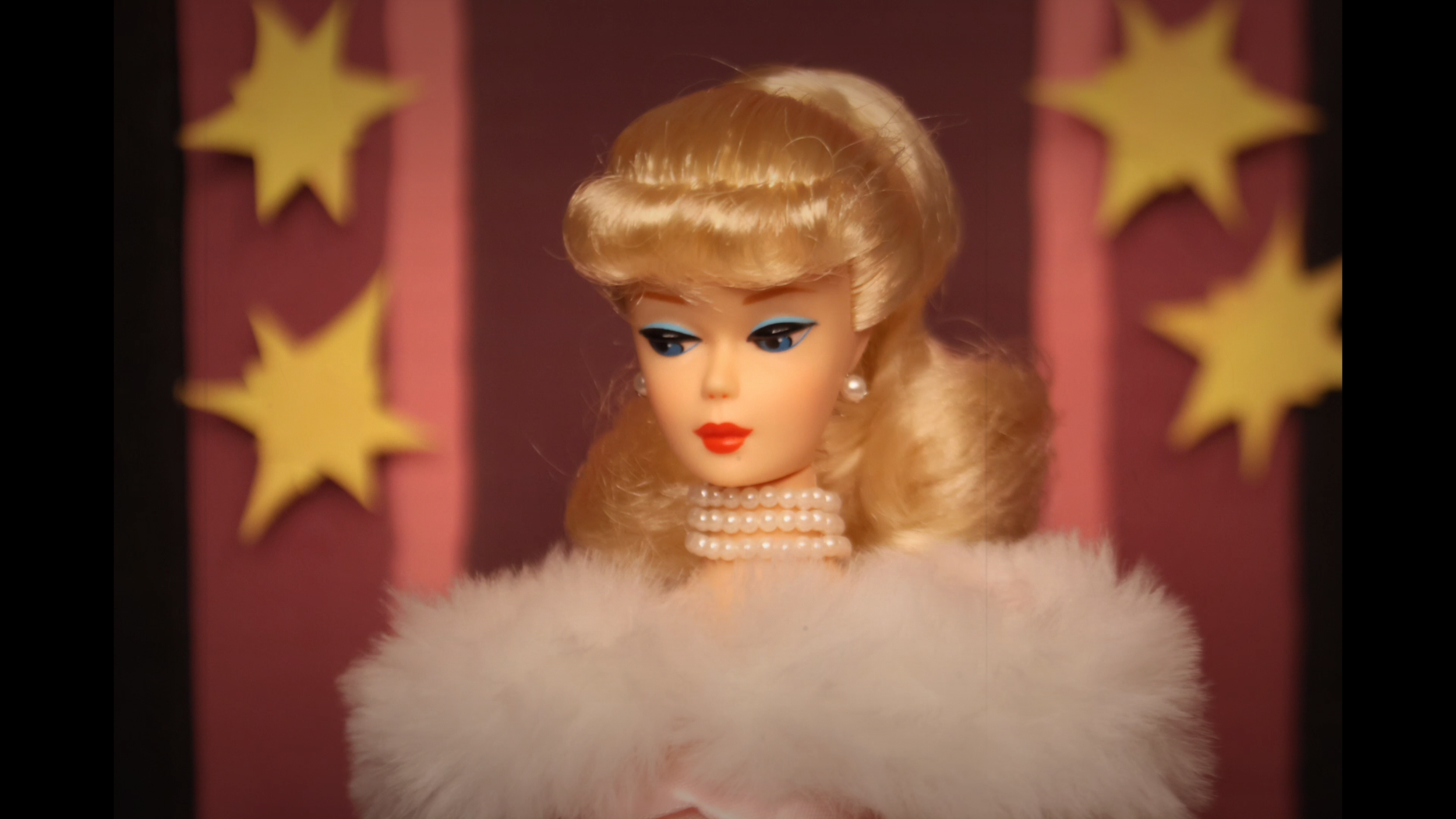 A vintage fashion doll with blonde hair styled in loose curls, wearing pearl earrings, a pearl necklace, and a faux fur stole, against a pink backdrop with yellow star shapes.