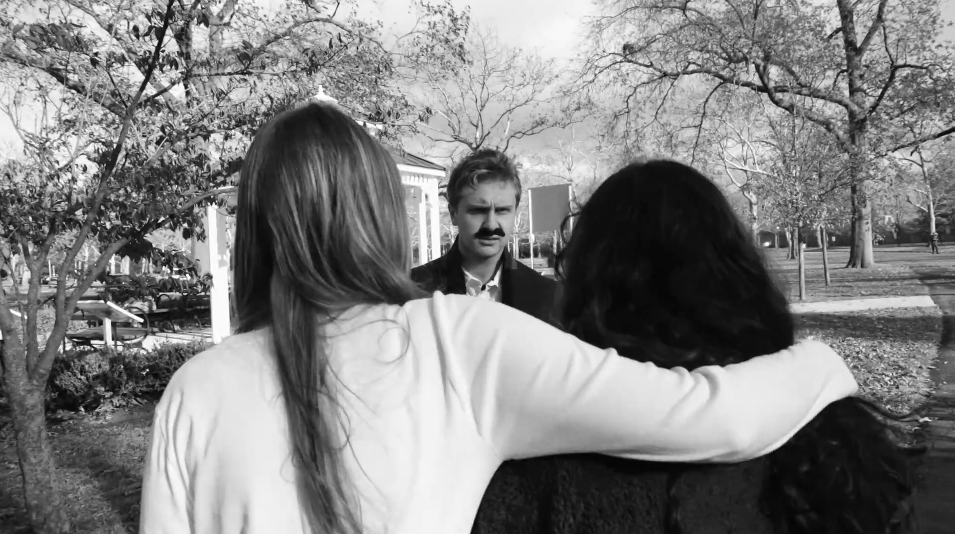 Two women with light-colored hair embracing with their backs facing the camera, and a man with light hair and a mustache standing in front of them, outdoors in a park with trees and a walkway.