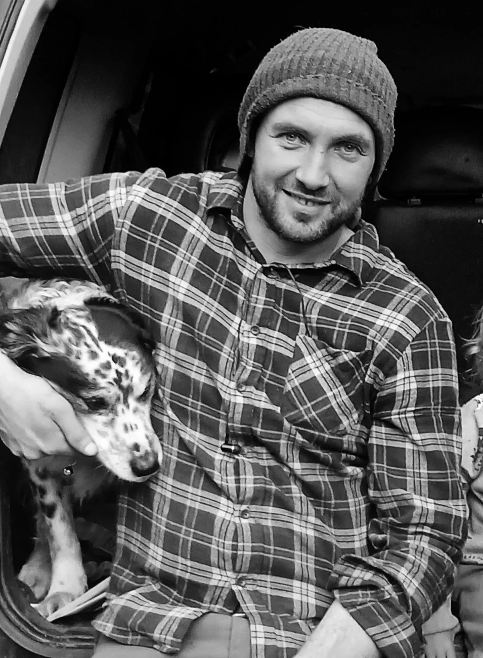 A man wearing a knit cap and a plaid shirt smiling while sitting next to a Dalmatian puppy.