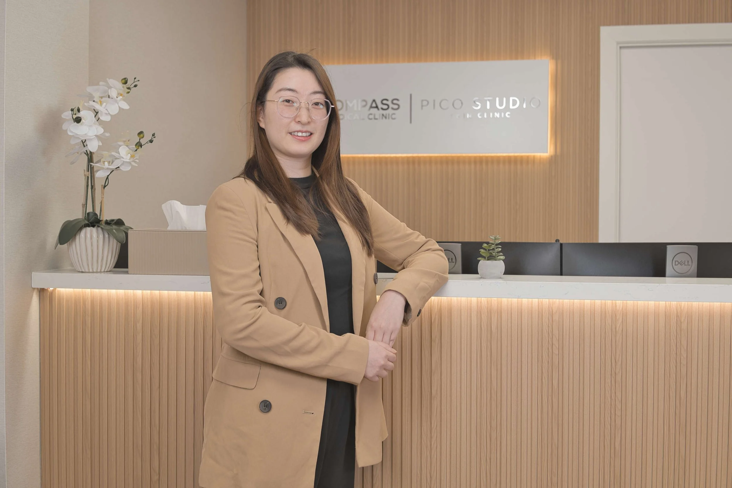 Woman wearing glasses and a tan blazer standing at a modern medical clinic reception desk