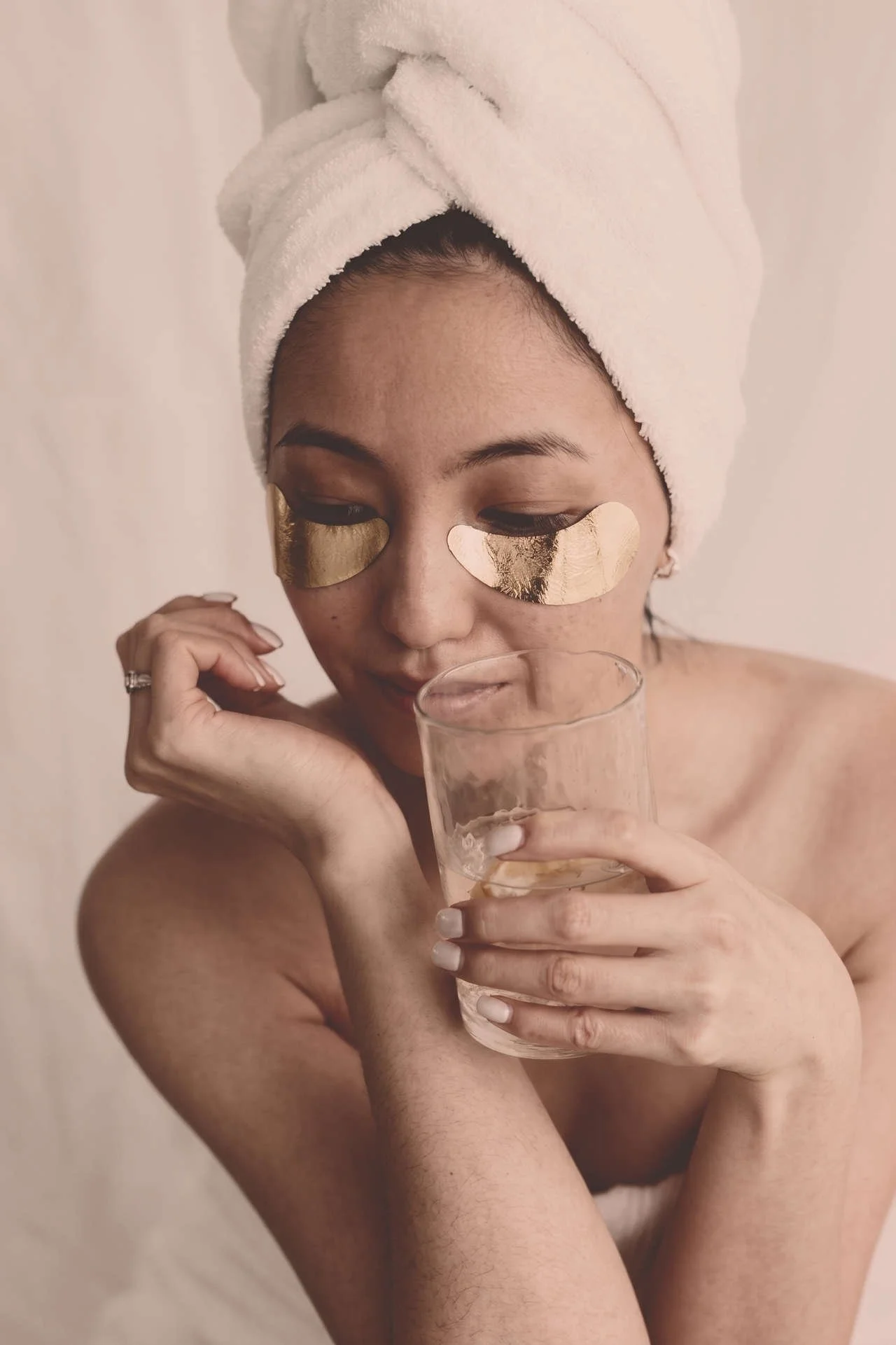 Woman wearing a towel turban and gold under-eye patches while holding a glass of water